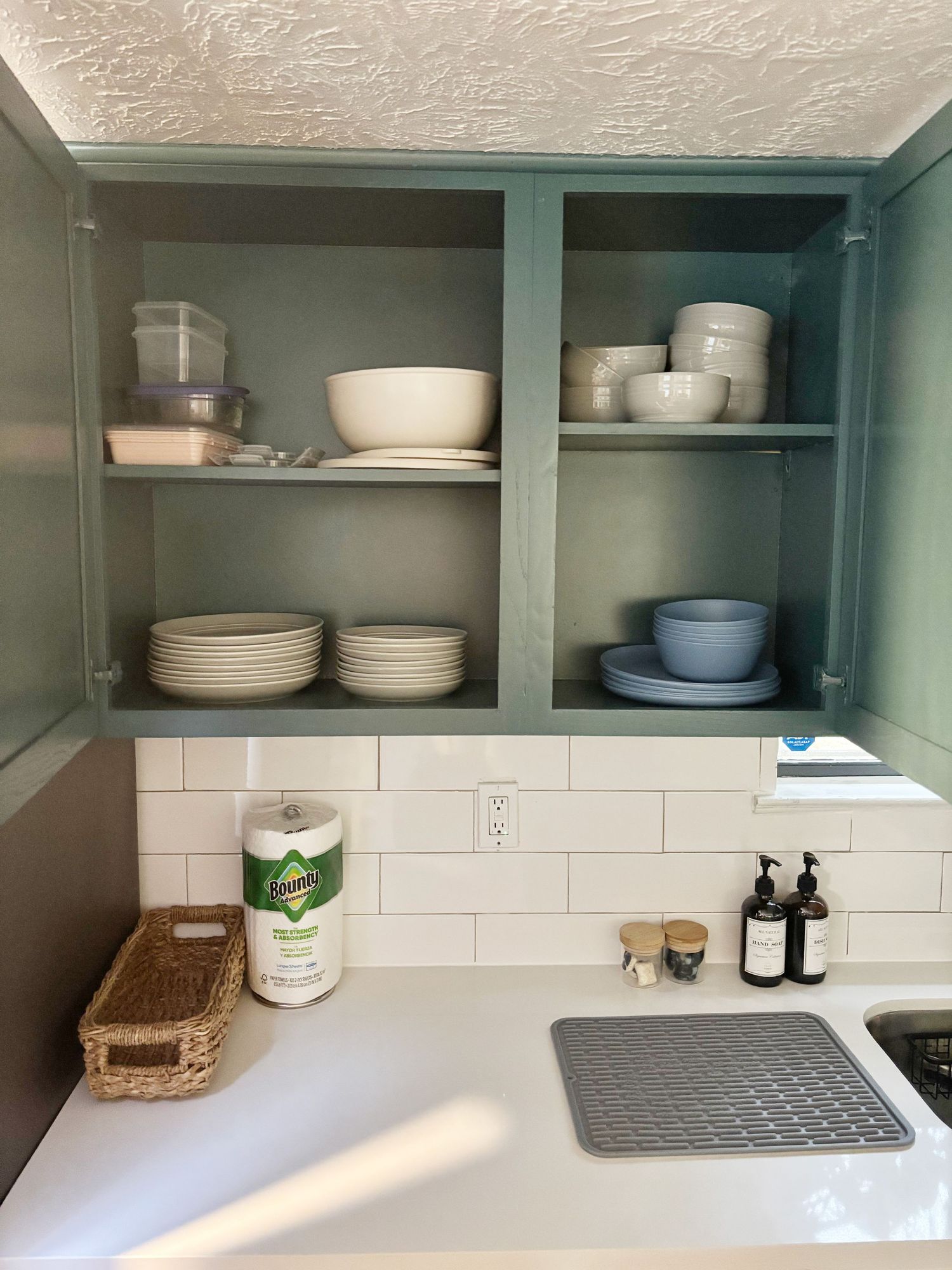 Sage green kitchen cabinets with white dishes, bowls, and plates on shelves above a white subway tile backsplash.