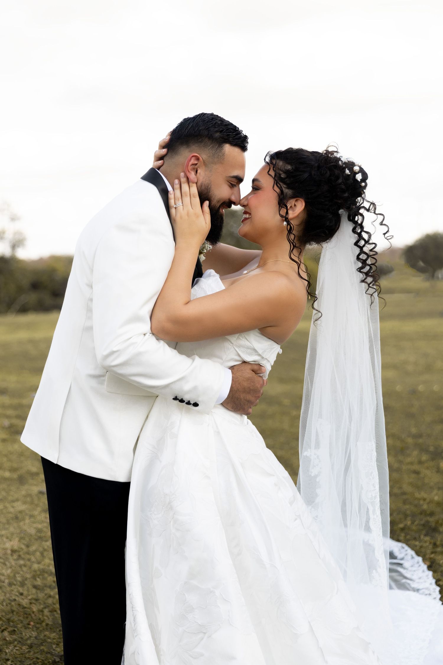 A romantic wedding couple embraces in a grassy field as the bride's veil flows in the breeze.