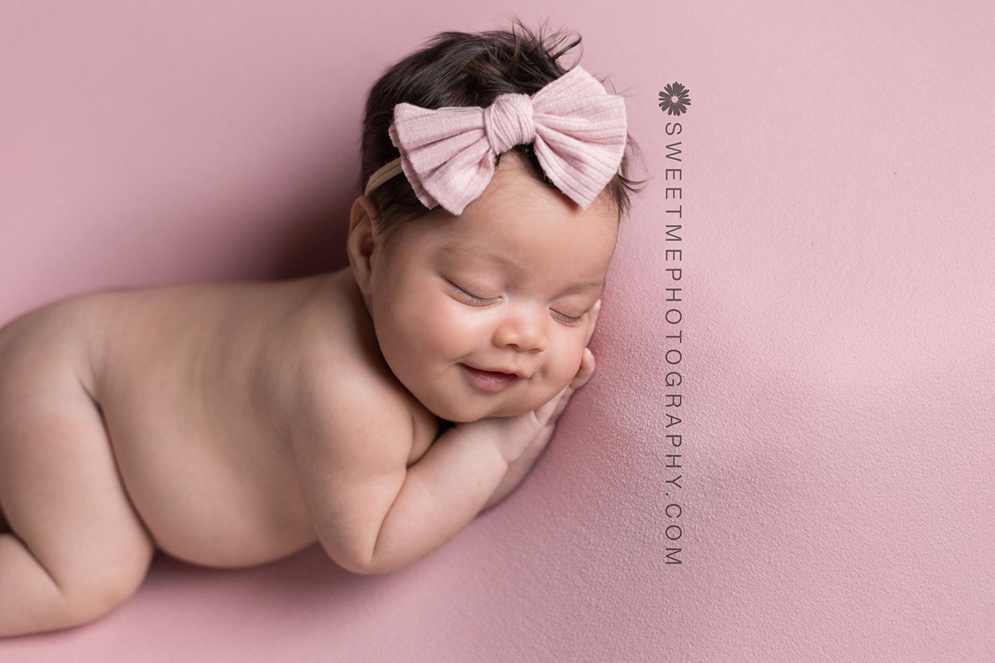 Sleeping newborn baby wearing pink bow headband on pink background in artistic studio portrait.