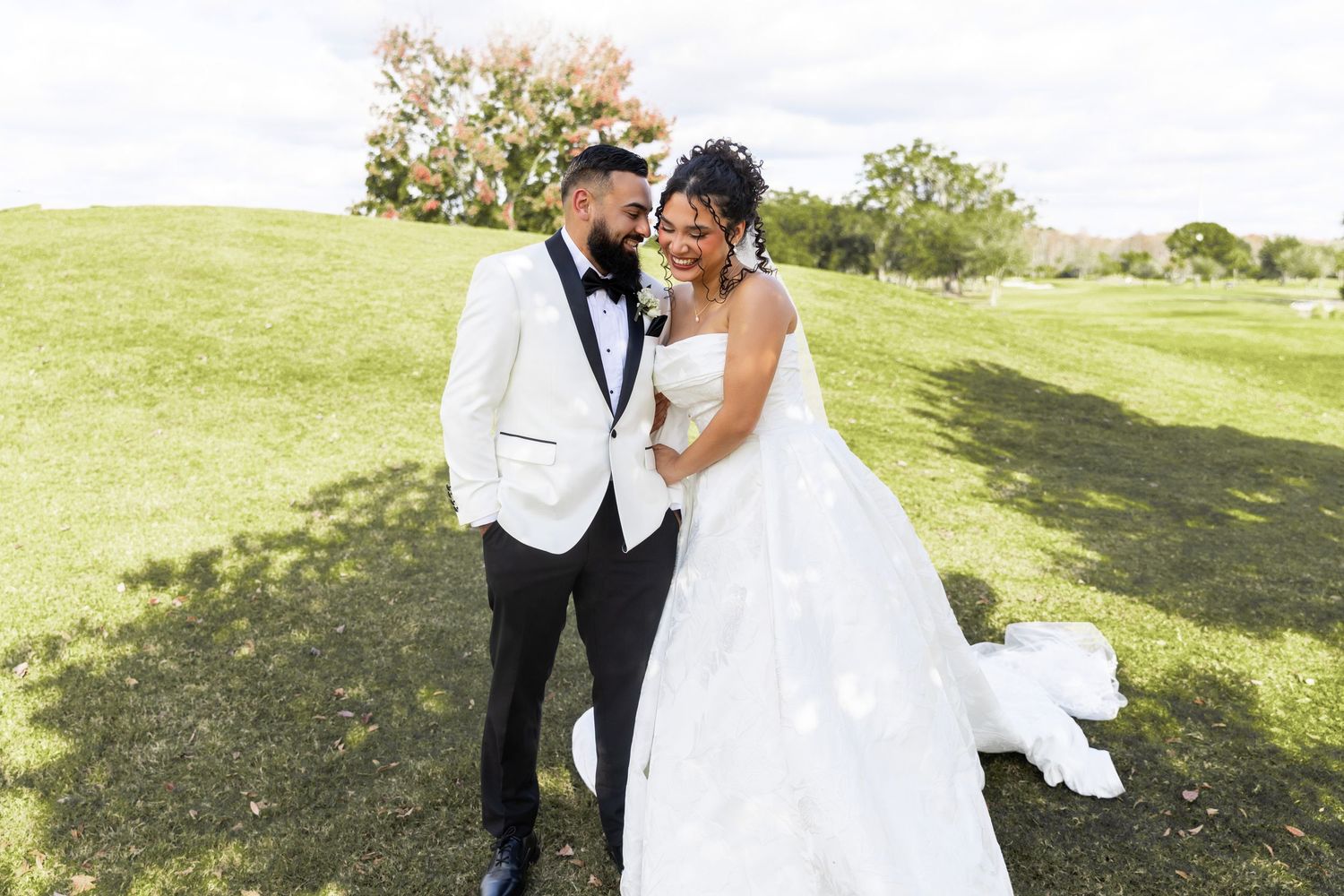 A couple in wedding attire embraces while dancing outdoors on a sunny day in a grassy field.