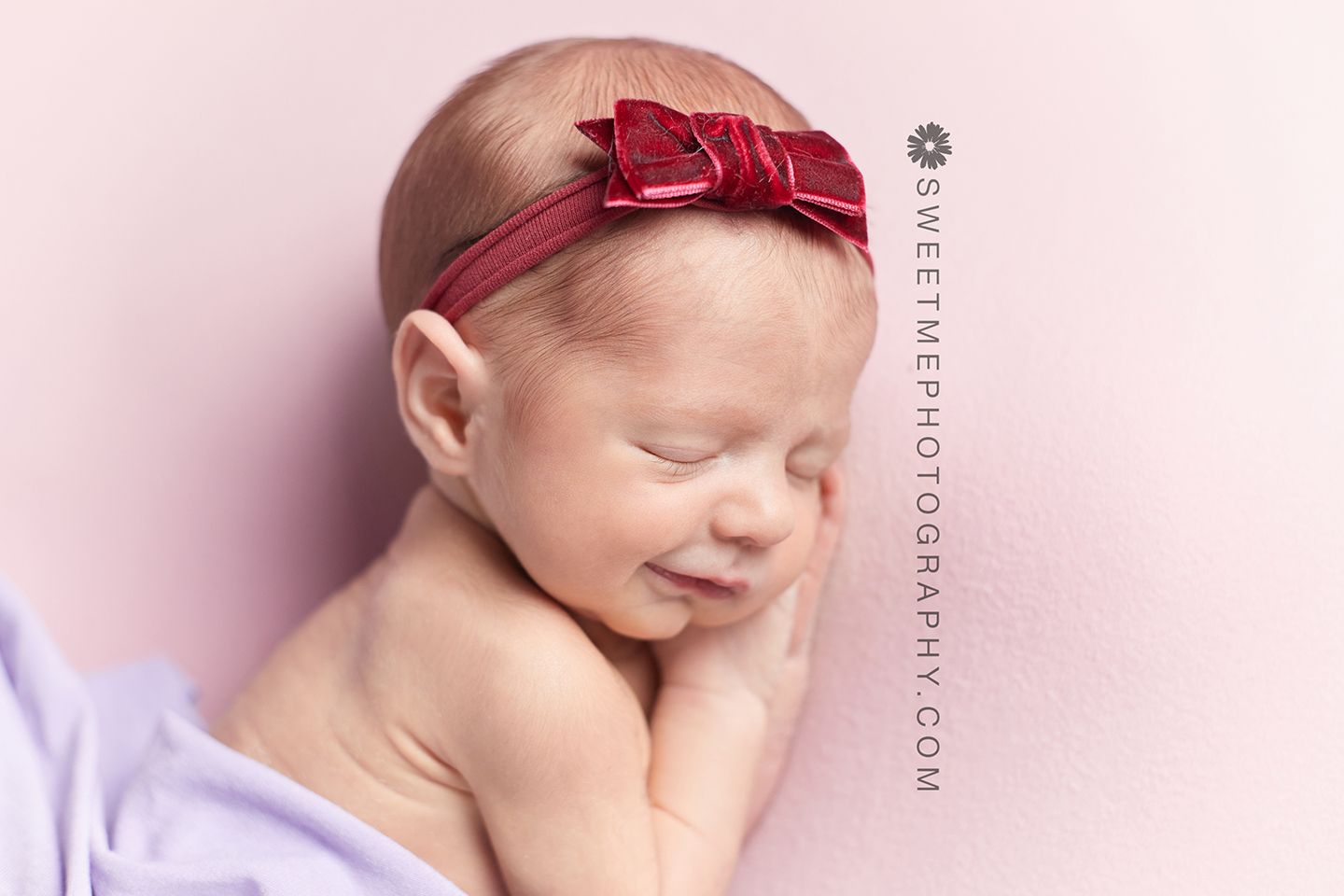 Sleeping newborn wearing red bow headband on pink background during professional photography session.