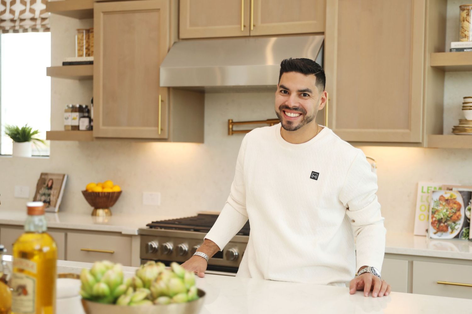 A modern white kitchen with glass cabinets and stainless steel appliances features someone preparing food at the counter.