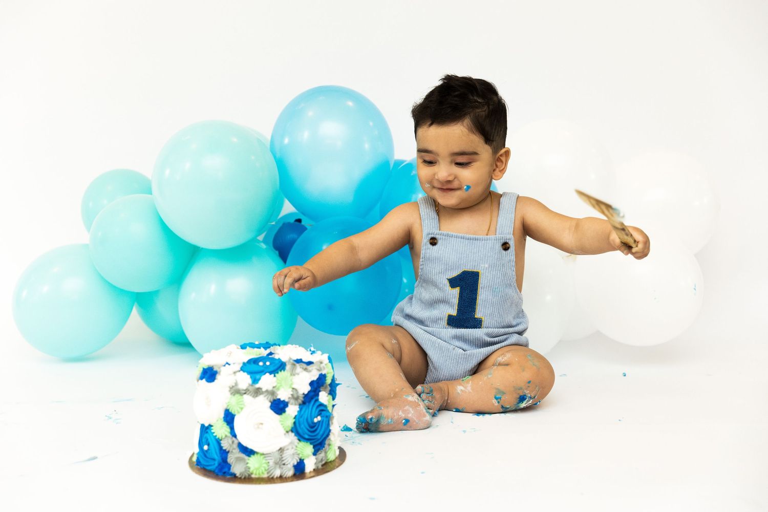 Baby boy in blue romper smashes first birthday cake surrounded by teal and white balloons on white background.