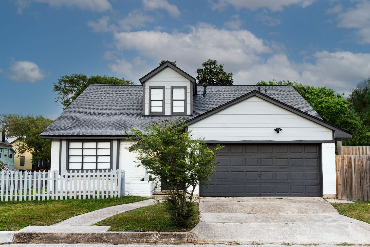 White Cape Cod home with gray roof, dormer window, dark garage door, and white picket fence on sunny day.