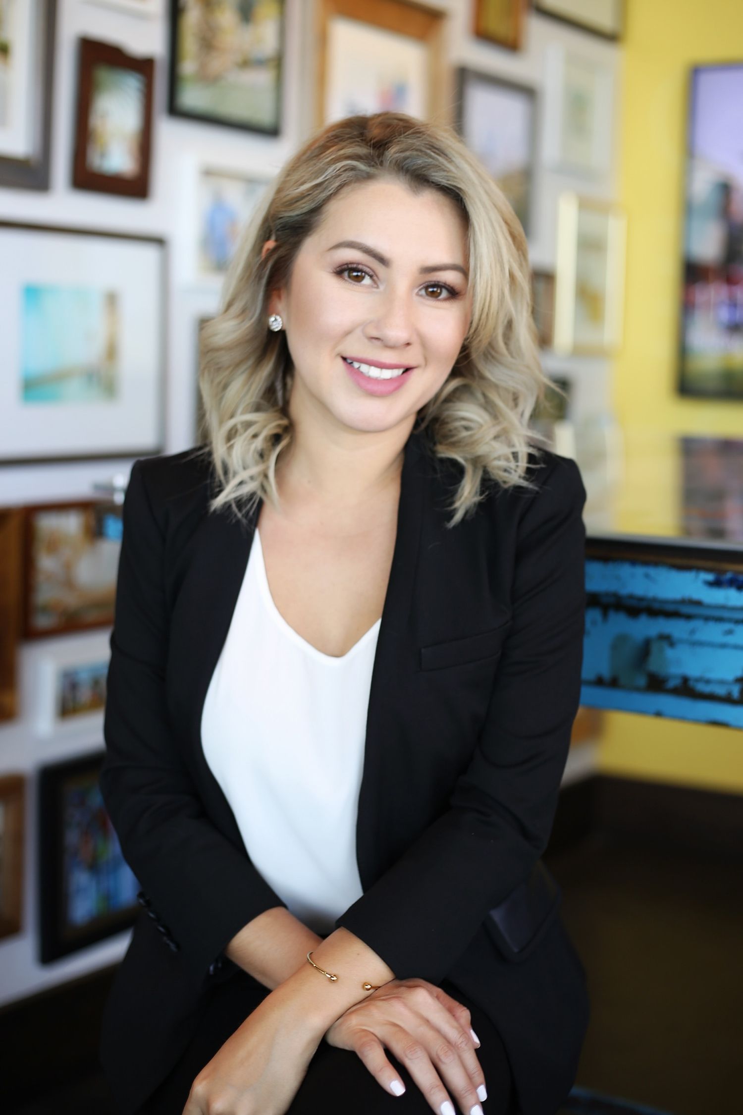 Professional headshot series of a person in a black blazer and white top against a yellow wall with framed artwork.