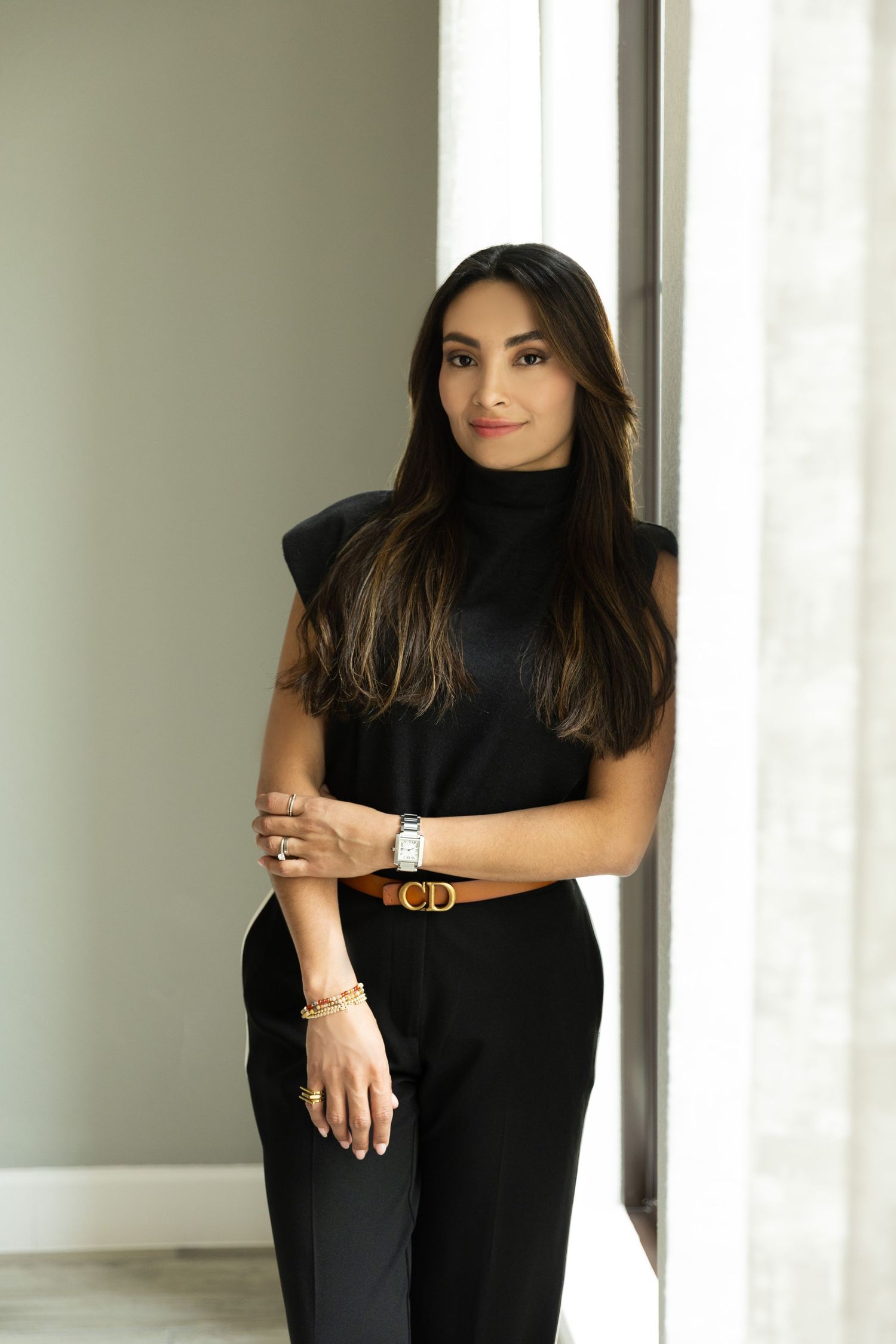 Stylish woman in black turtleneck jumpsuit with brown Dior belt and watch, posing elegantly indoors.