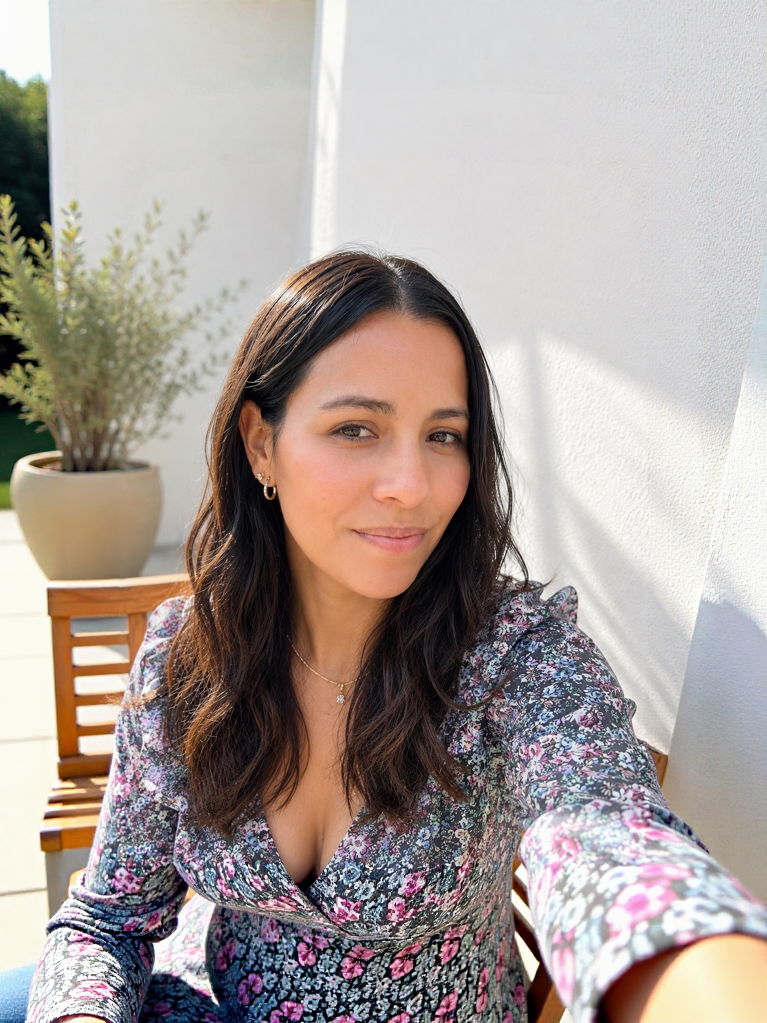 Woman in floral dress taking a selfie outdoors on a sunny patio with potted plants in the background.