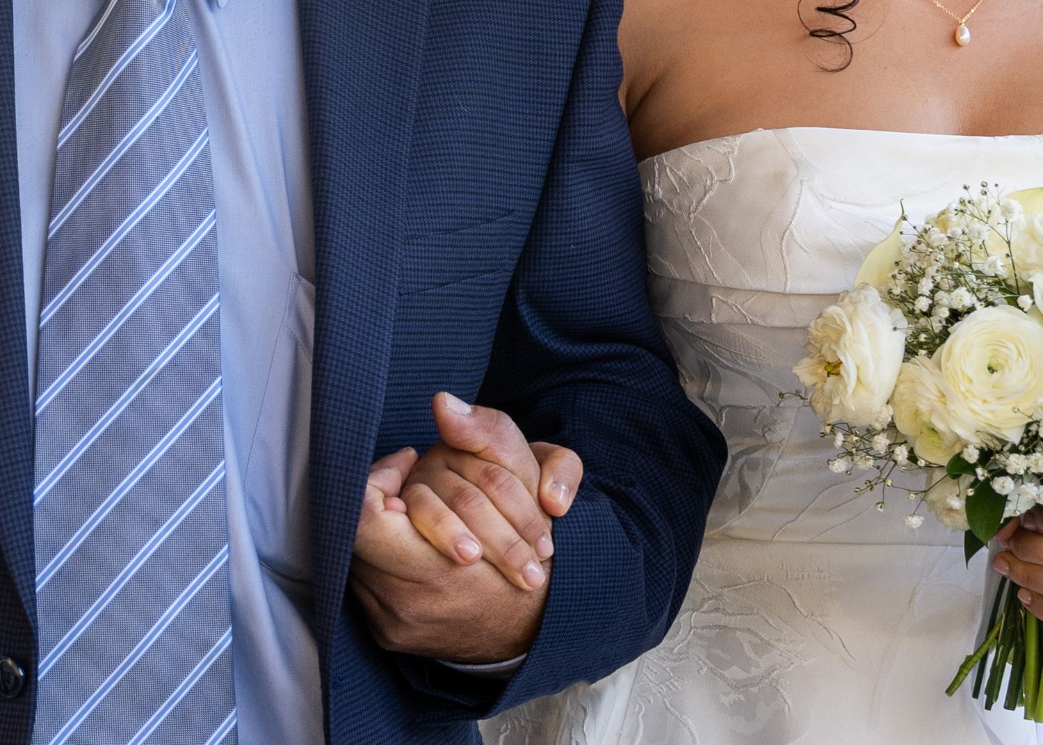 Close-up detail of wedding couple holding hands while wearing formal navy suit and white lace dress with bouquet.