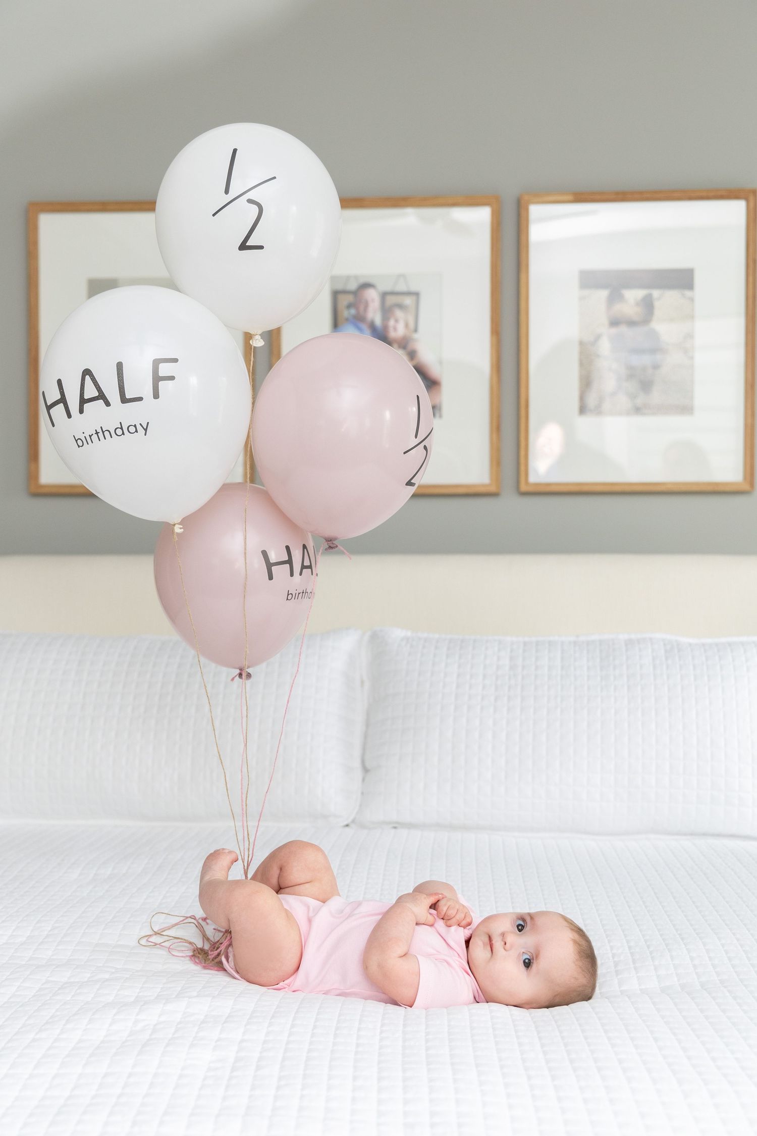 Baby girl in pink onesie lying on white bed surrounded by half birthday balloons for 6 month milestone photo.