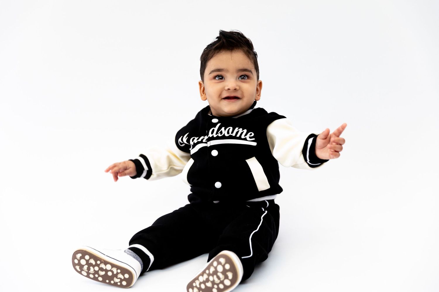 Cute baby boy sitting in a black varsity jacket with Handsome text, wearing matching pants and shoes on white background.
