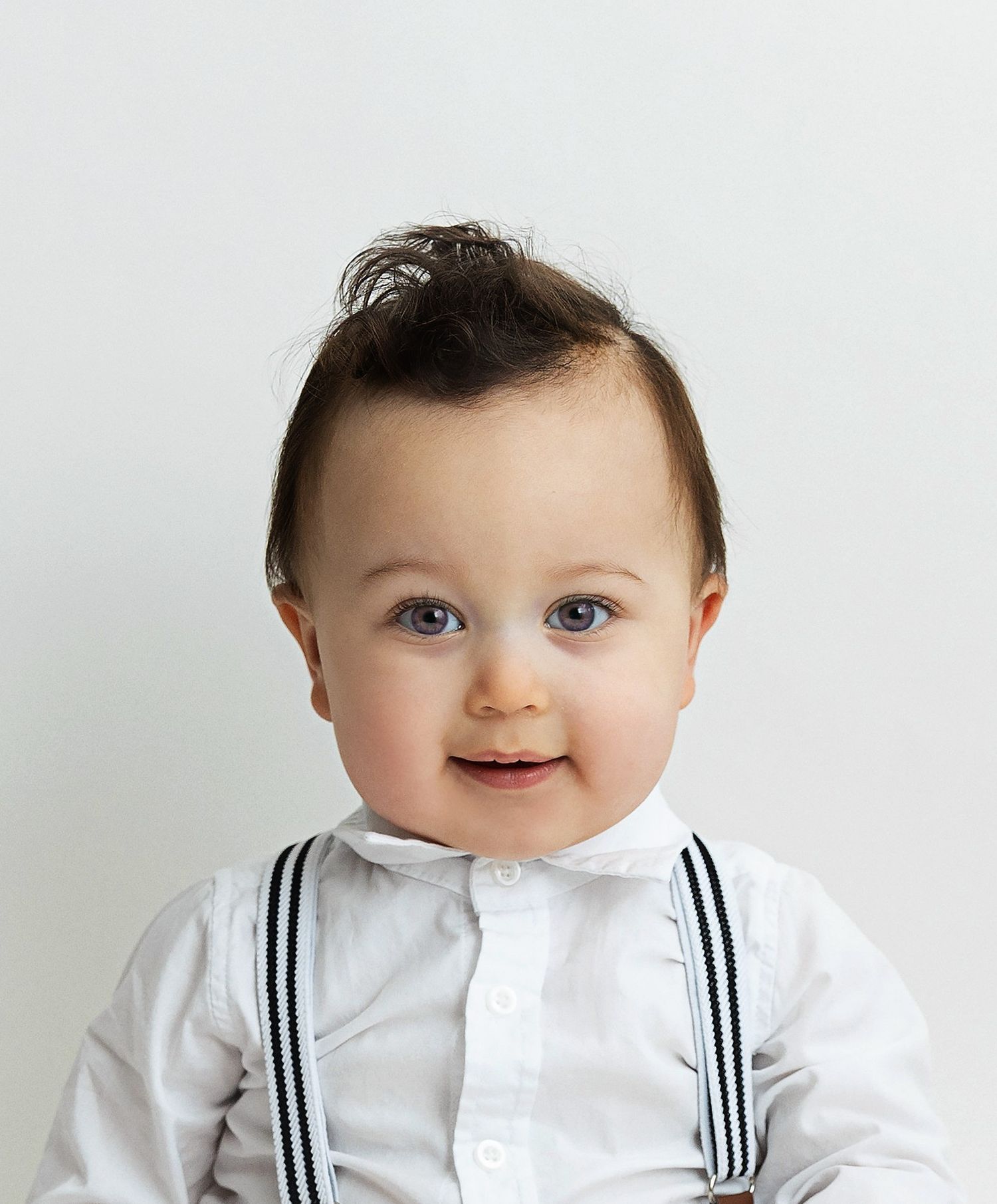 A baby wearing a white shirt with suspenders smiles while making different playful expressions.