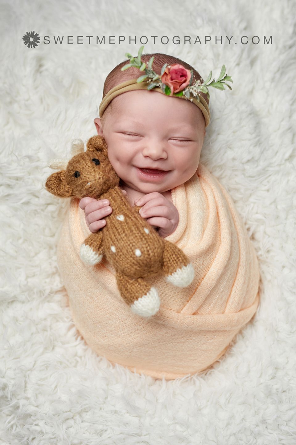 Newborn wrapped in peach blanket holding teddy bear on white fur with floral headband.
