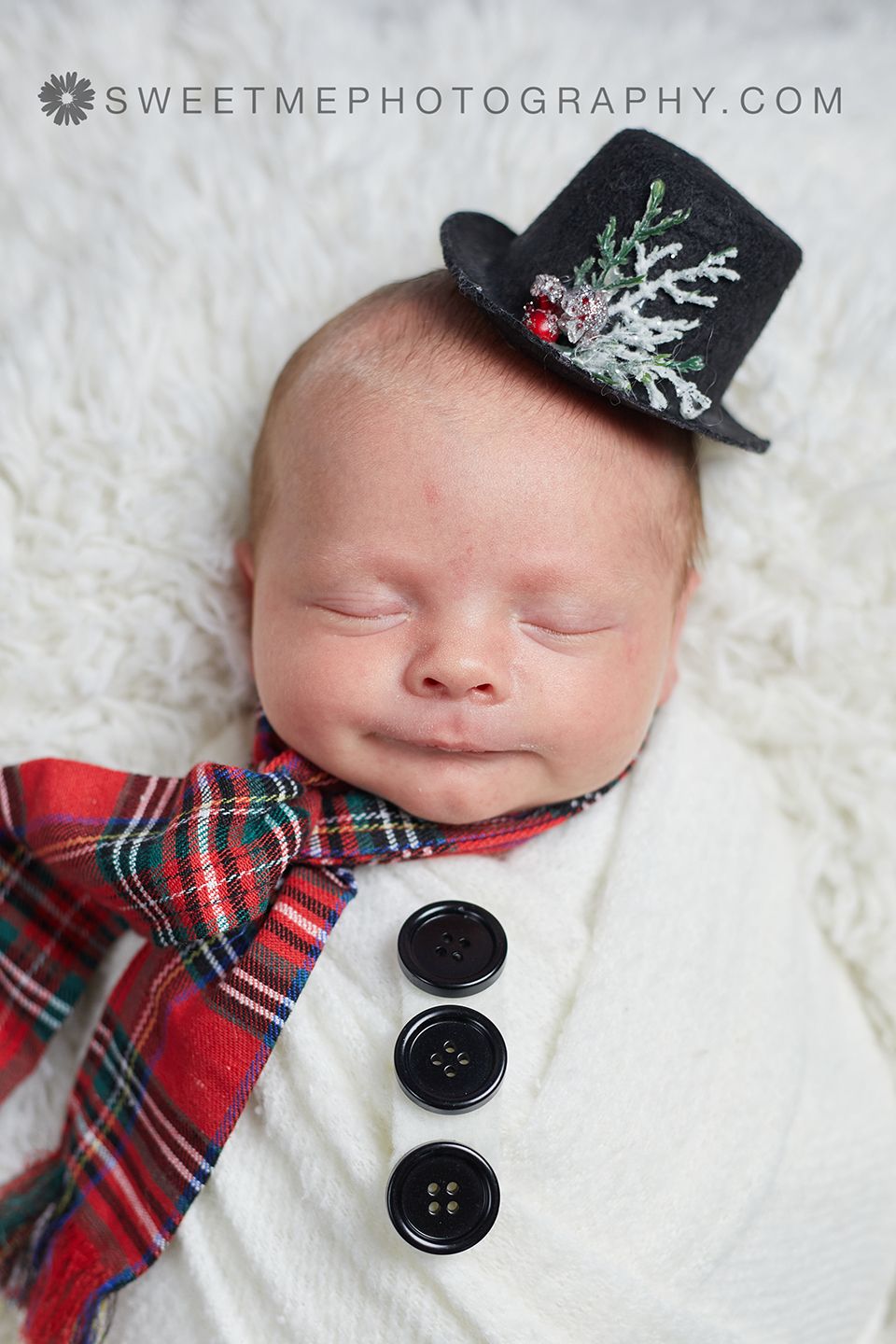 Newborn wearing top hat and plaid bowtie sleeping on white fur blanket with decorative buttons.