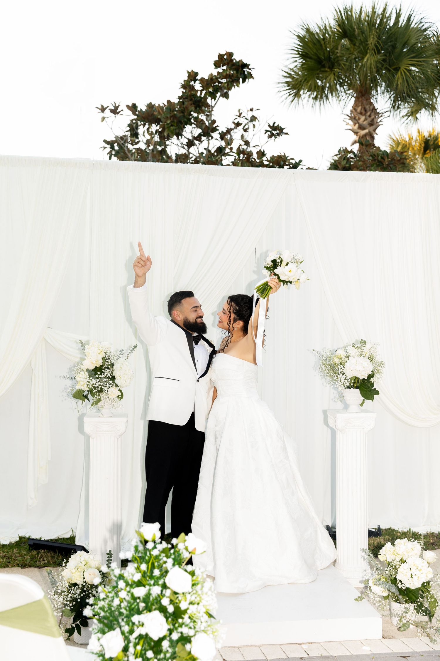 Elegant white wedding backdrop with floral arrangements and flowing fabric panels creates a romantic outdoor ceremony setting.