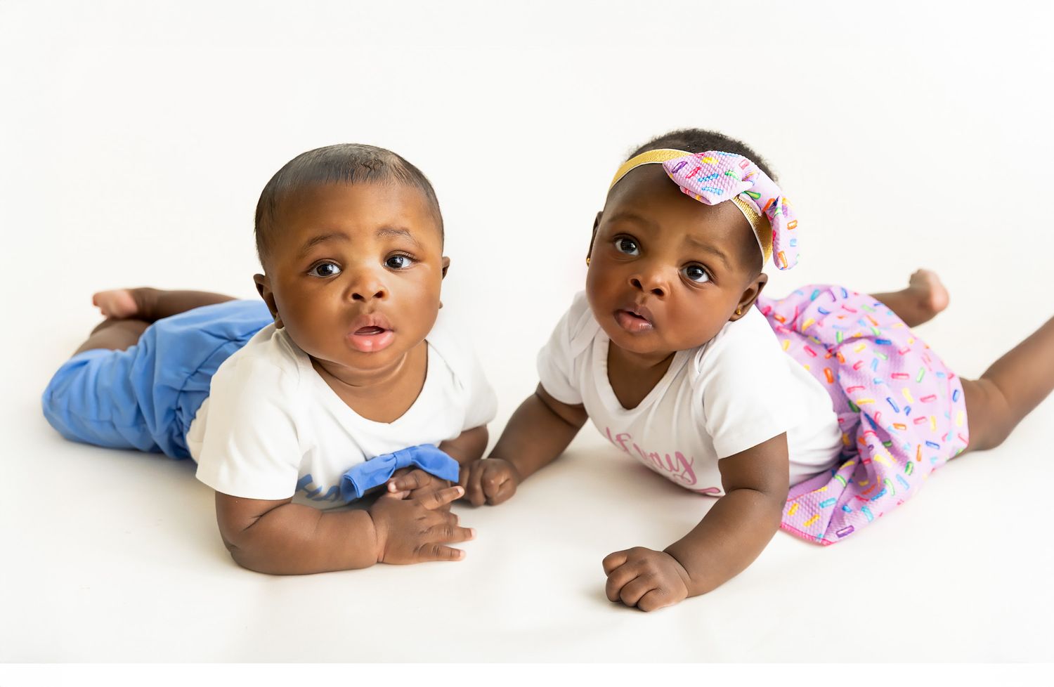 Two adorable Black babies in white shirts lying on their stomachs, one in jeans and one in a pink skirt with headband.