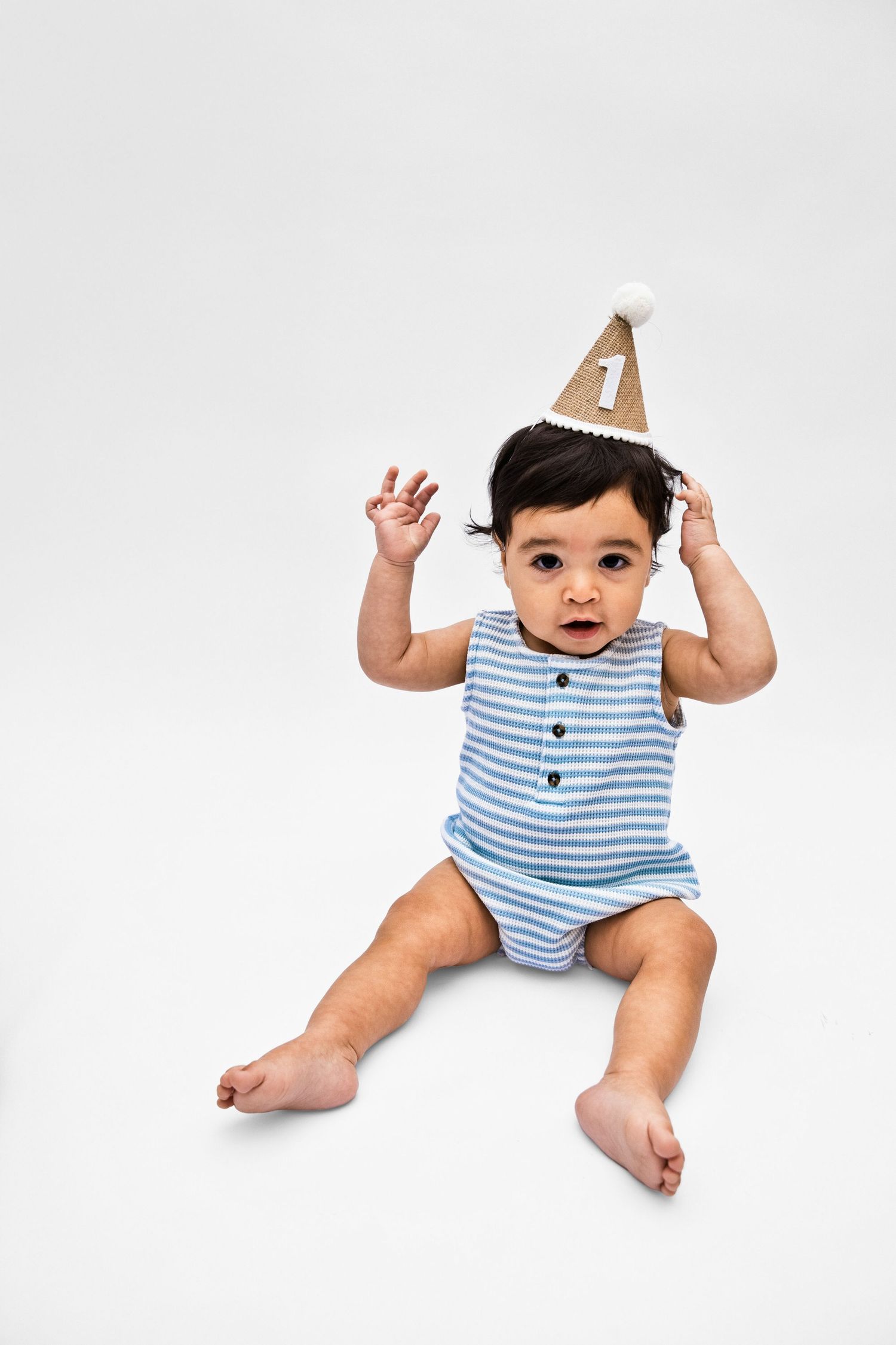 Cute baby in blue striped onesie wearing a number one birthday party hat, sitting on white background.