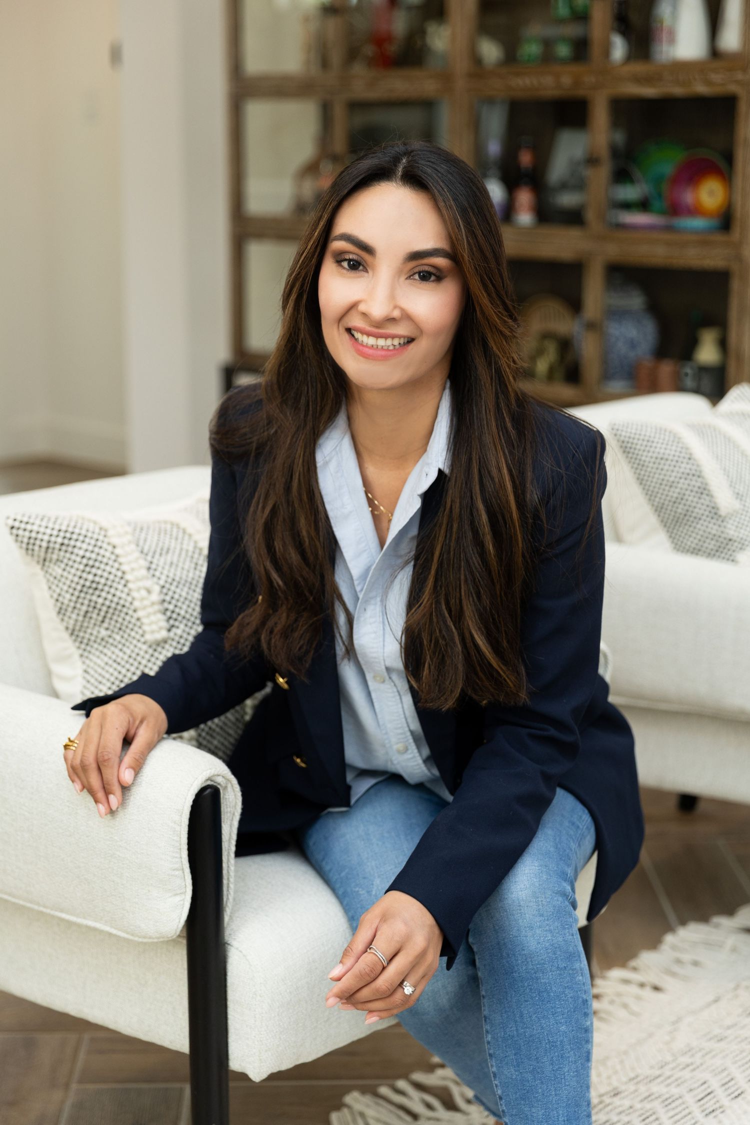 A smiling woman with long dark hair sits on a white sofa wearing a navy blazer and jeans in a modern living room.