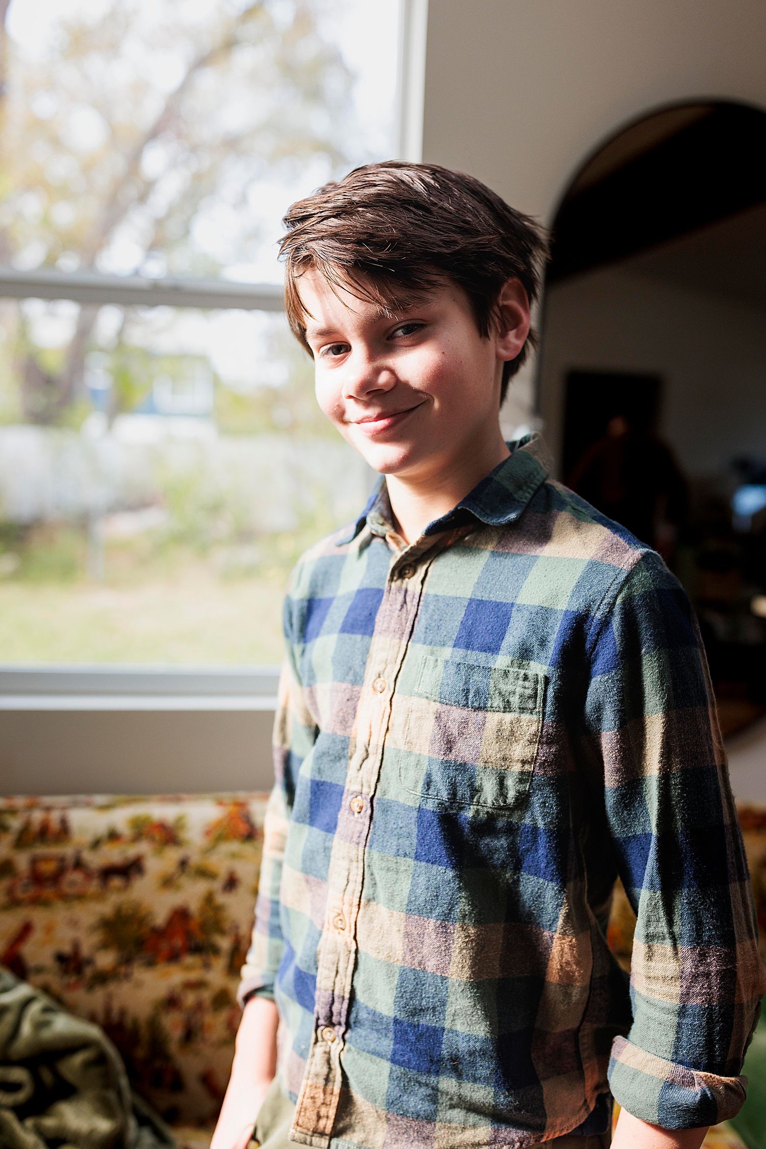 son stands in front of window in plaid shirt for lifestyle session in studio