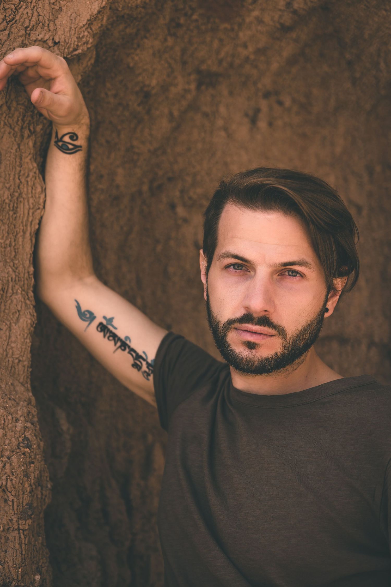 Man with short hair and arm tattoos posing against a textured rock wall of rock, gazing directly at the camera, exuding earthy, edgy tones.