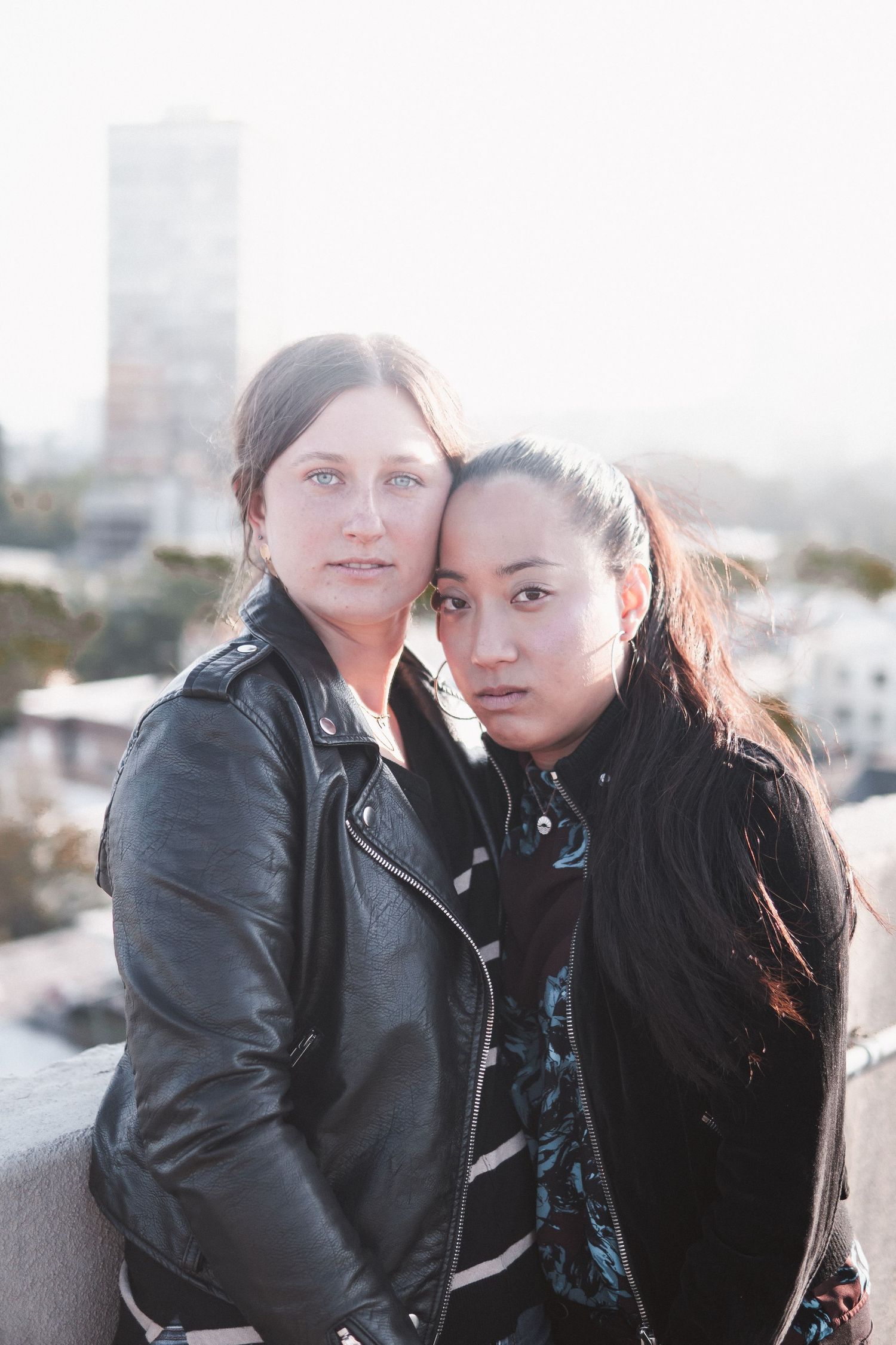 Soft minimal portrait of two women standing close on a rooftop, captured in natural light with an urban backdrop and calm, intimate energy