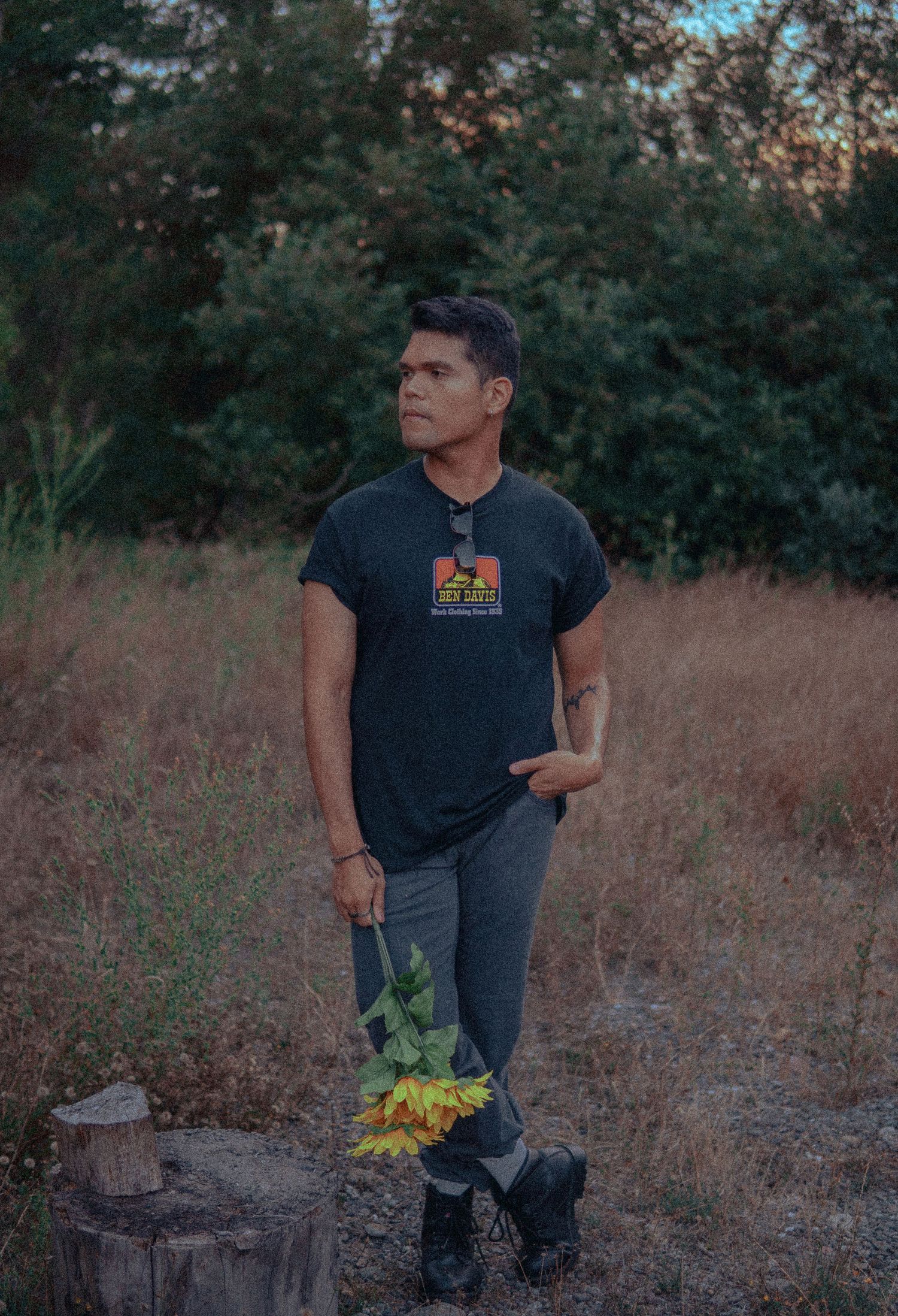 Man standing in a field at dusk holding sunflowers, wearing a black T-shirt and boots.