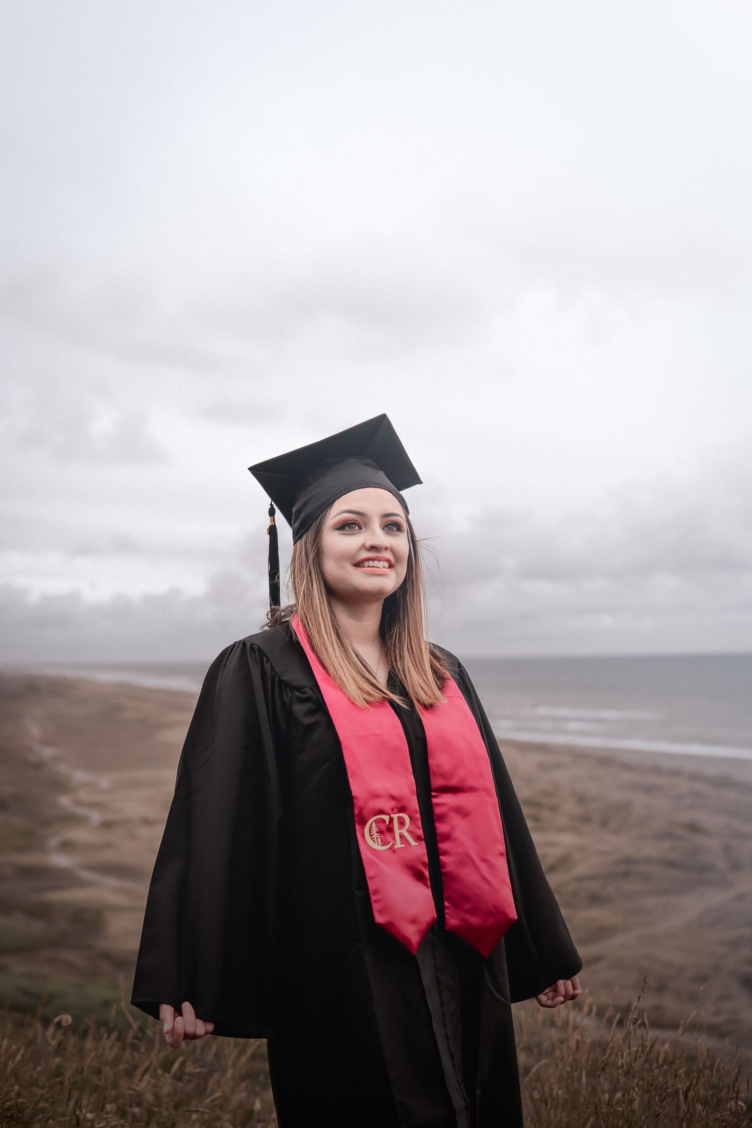 Outdoor graduation photo of a woman in cap and gown standing on a coastal bluff with cloudy skies and ocean views behind her.