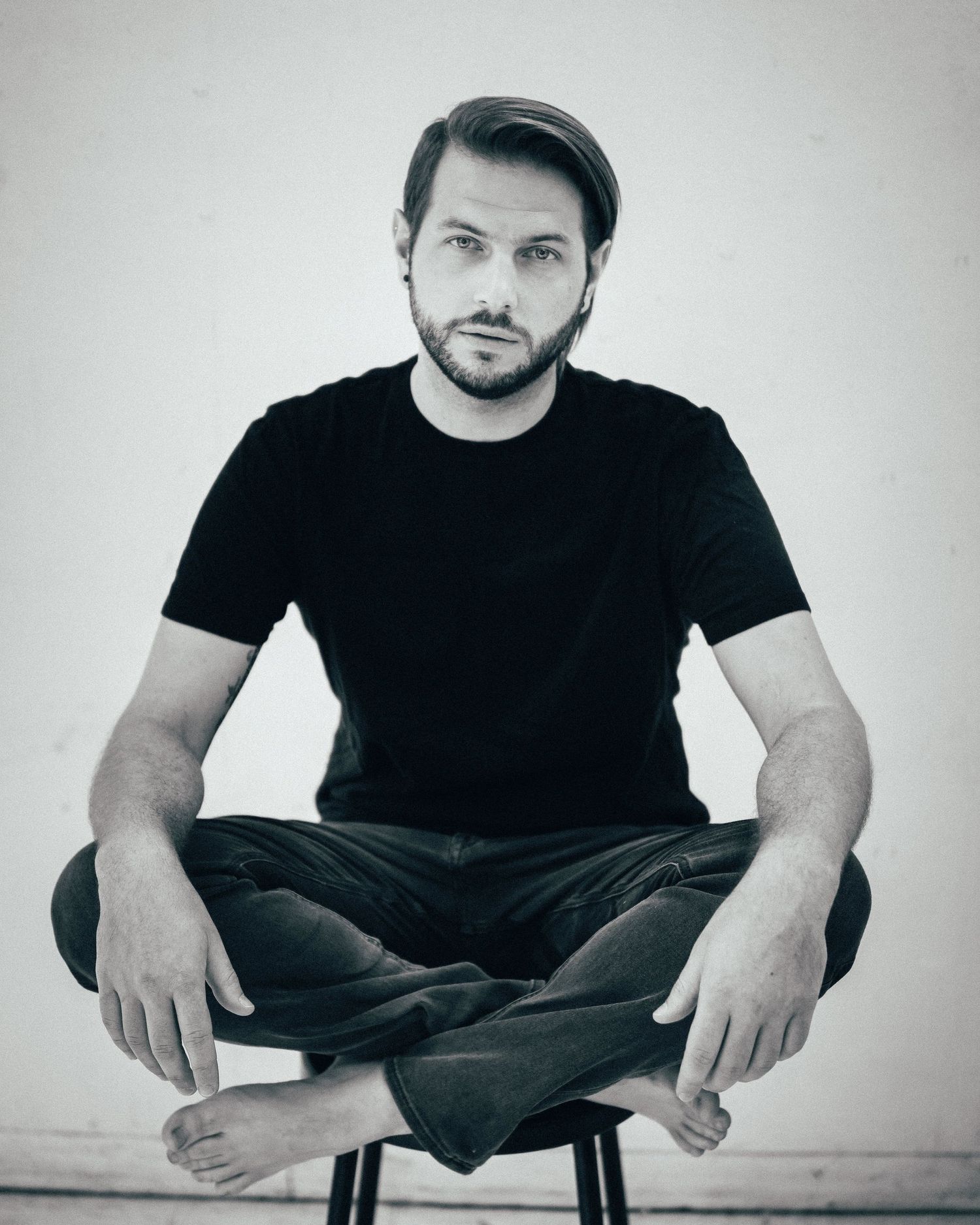 Minimalist black and white portrait of a man wearing a black t-shirt, sitting cross-legged on a stool, indoors with soft, natural lighting .