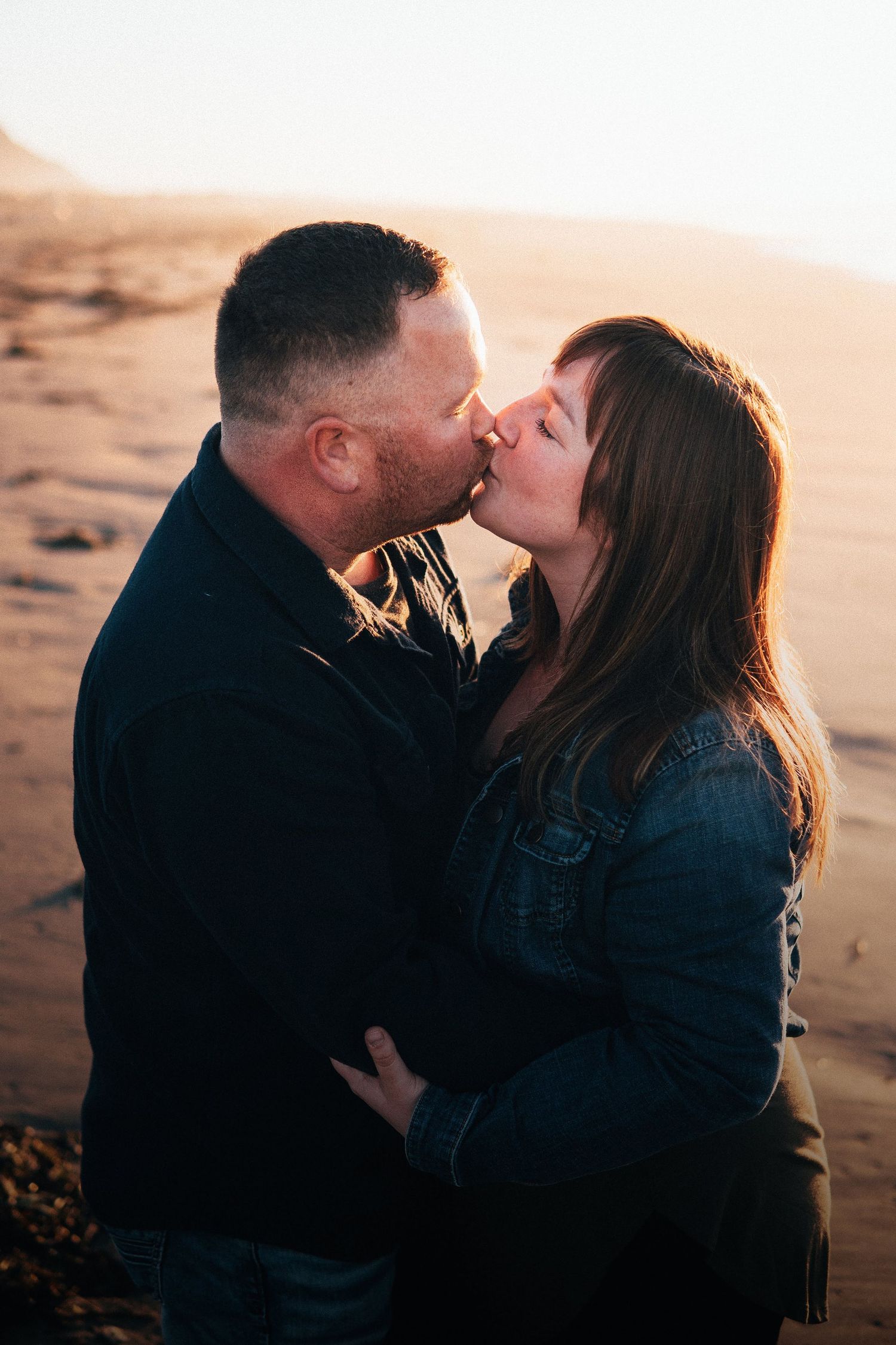 Couple sharing a kiss on a sandy beach at sunset, warm golden light highlighting their faces during a tender shoreline moment.