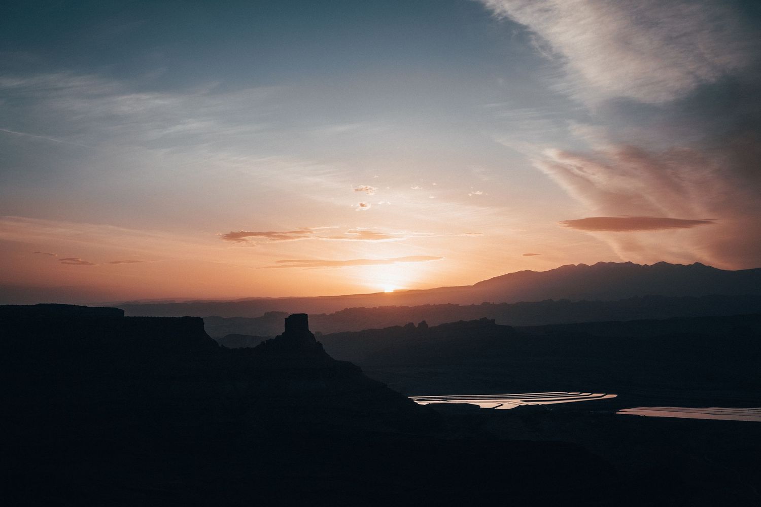 Golden sunrise over layered desert mesas with soft pastel skies and reflective water below, creating a calm, expansive adventure landscape.