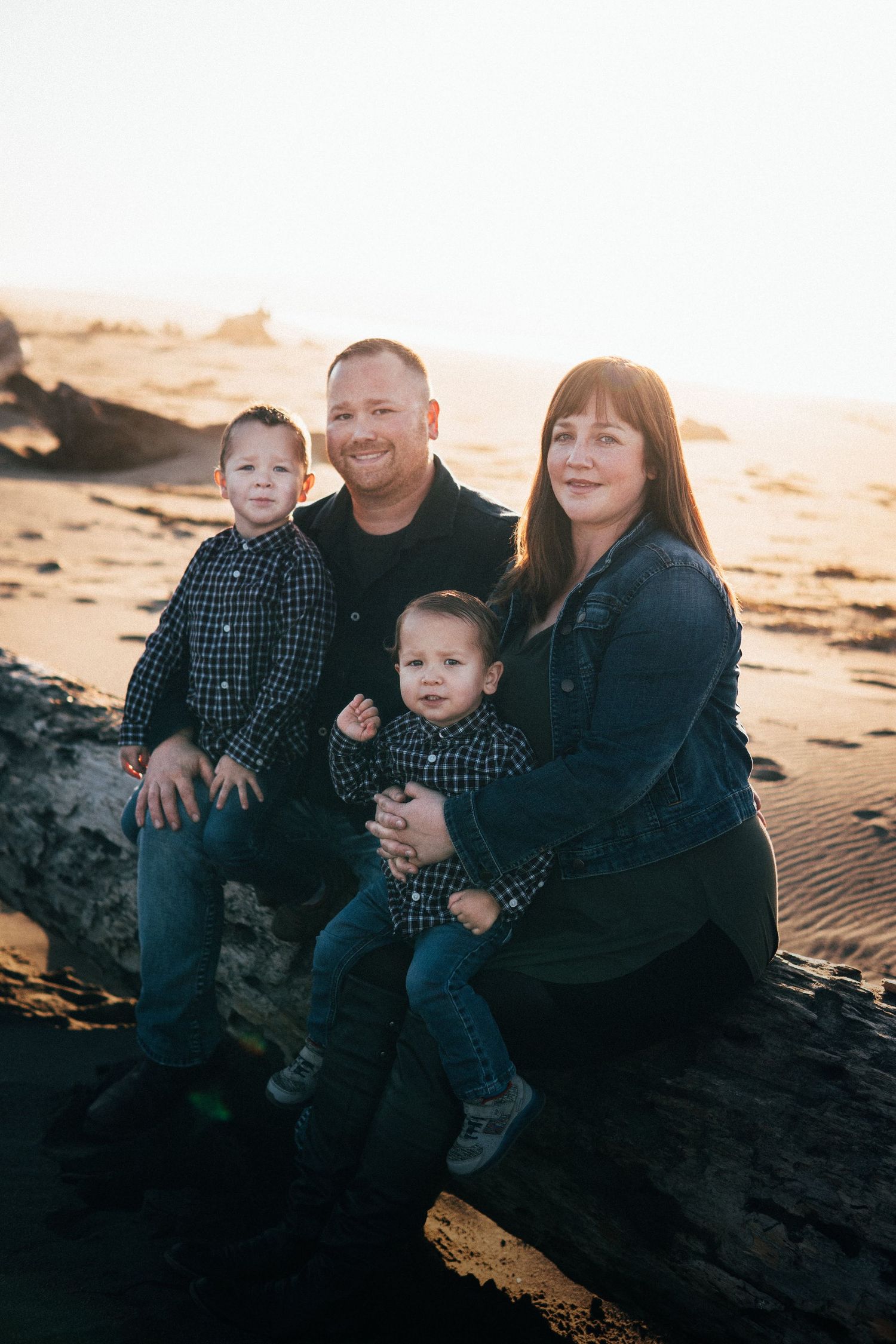 Family sitting on a driftwood log at the beach during golden hour, smiling together with warm sunlight and coastal scenery.