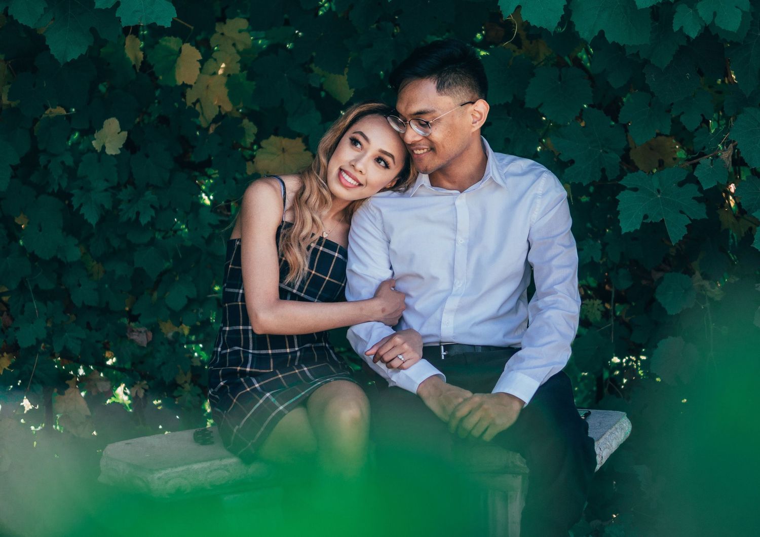 Couple sitting close on a stone bench surrounded by dense green leaves, sharing a warm, intimate moment in soft natural light