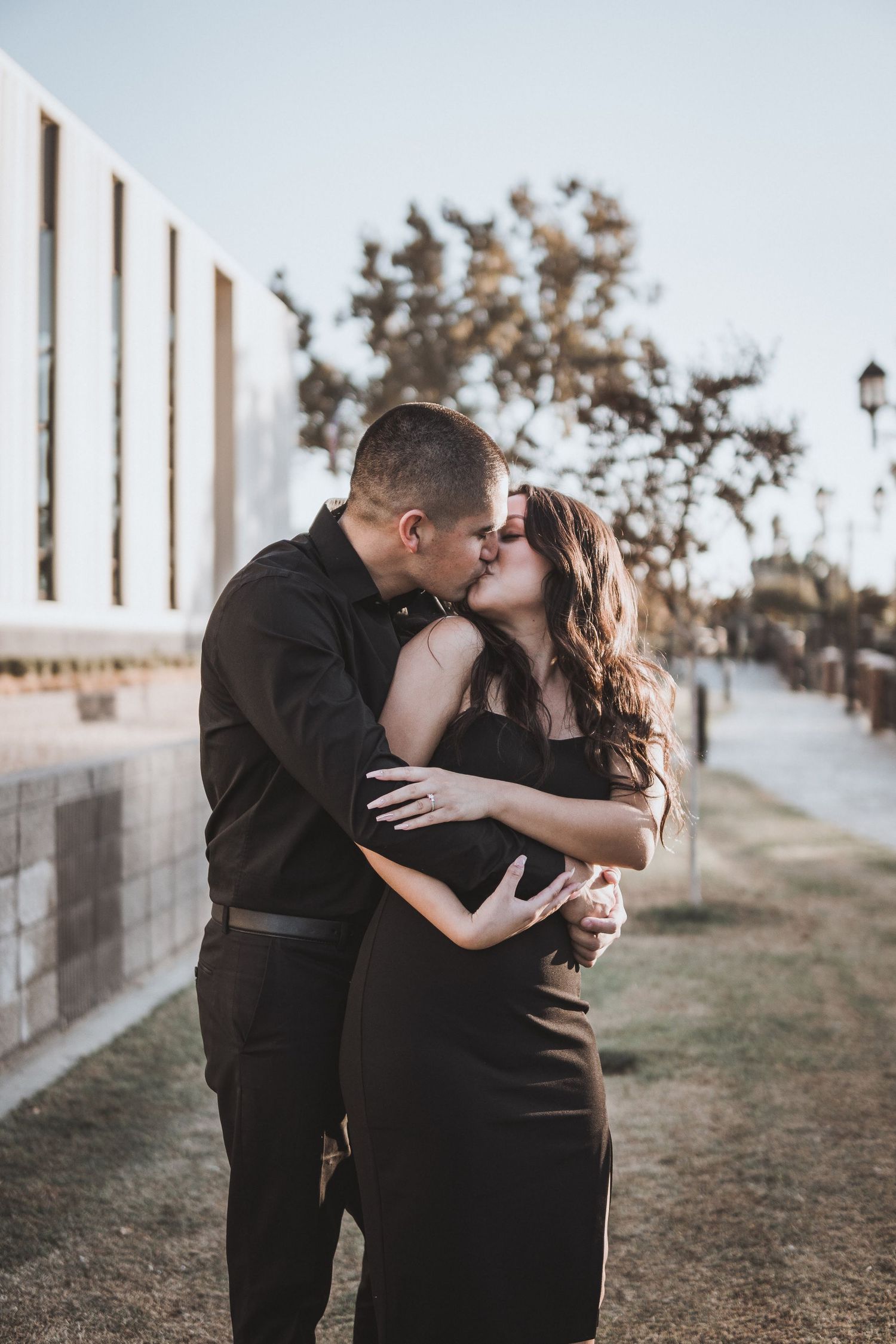 Couple embracing and kissing along a sunlit walkway near a modern building, with warm golden light creating a soft, romantic outdoor moment.