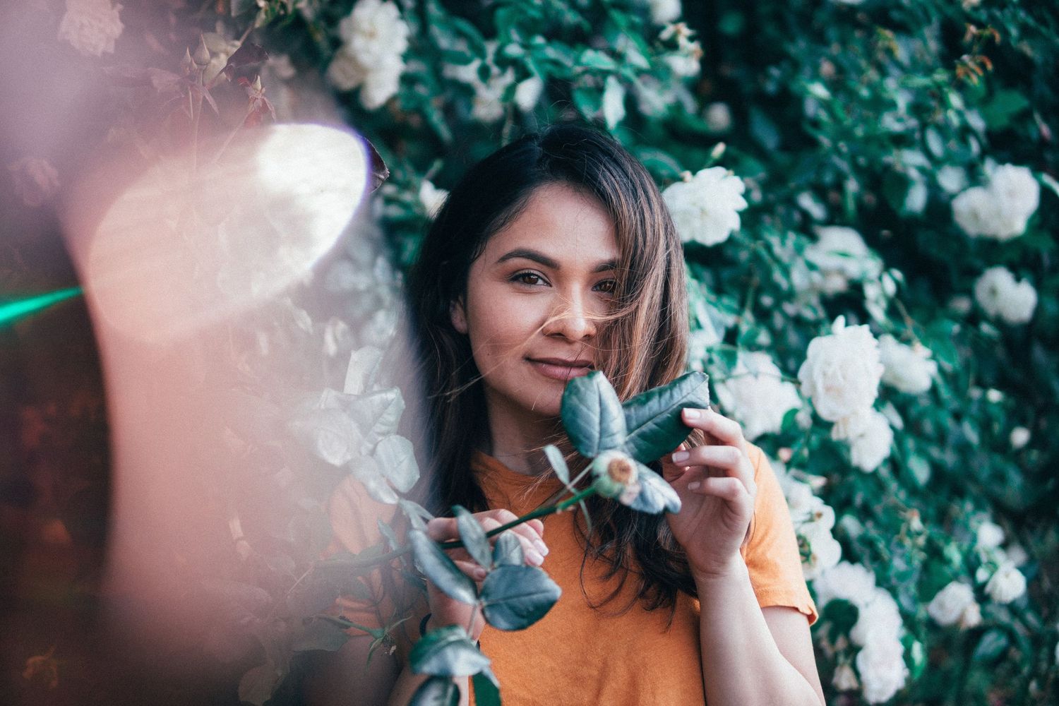 Woman standing among blooming white roses, smiling softly while holding leafy branches, creating a natural, serene portrait with flowers