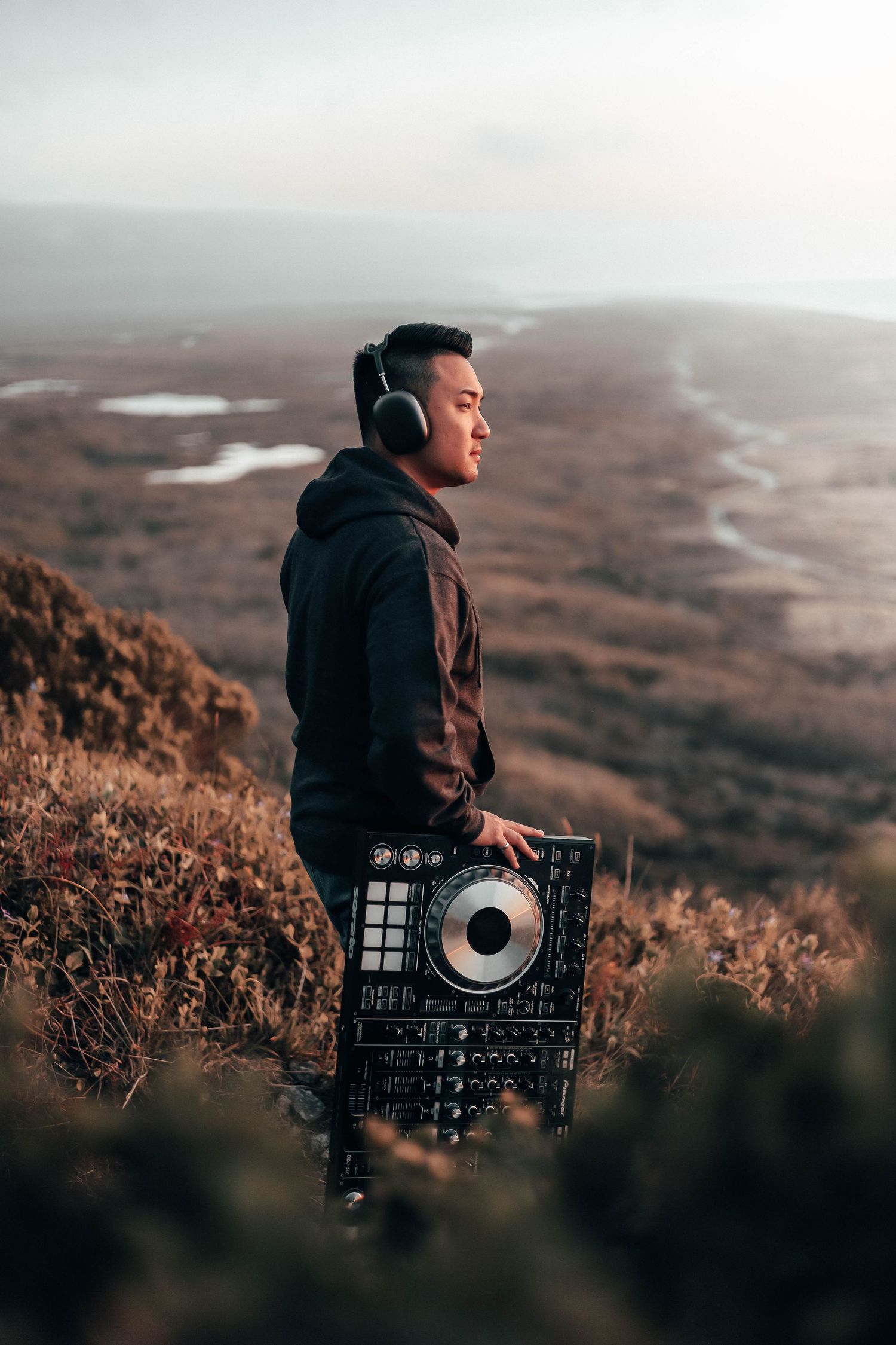 Person wearing headphones and holding DJ equipment on a cliffside at sunset, overlooking a wide valley and winding river landscape.