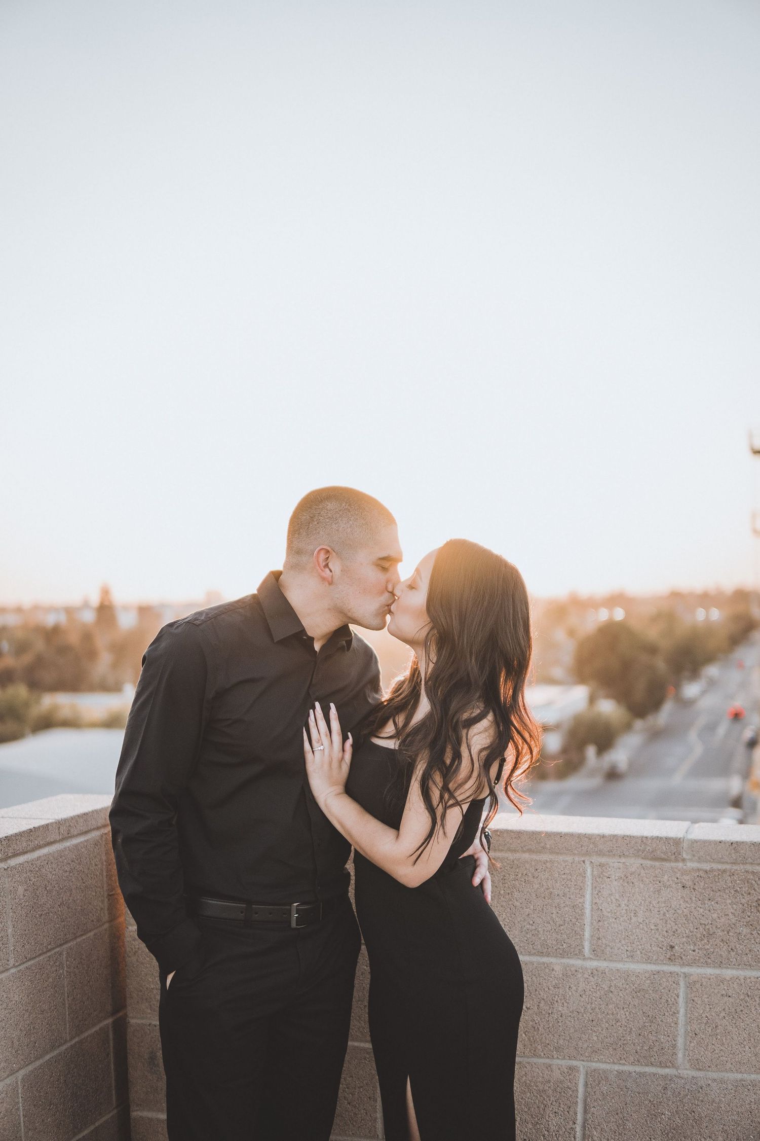 Romantic couple sharing a sunset kiss on a rooftop overlook, dressed in black with city views and warm evening light behind them.