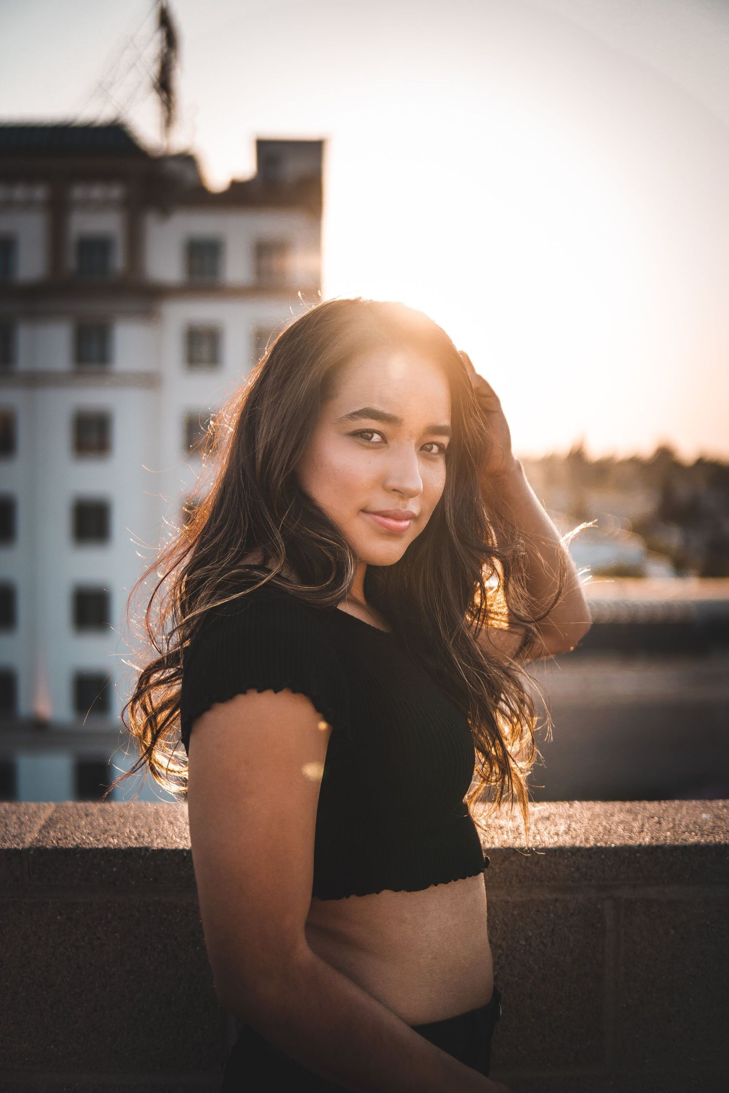 Golden-hour rooftop portrait of a young woman in a black top, glowing sunlight behind her, soft urban background, warm cinematic lighting.