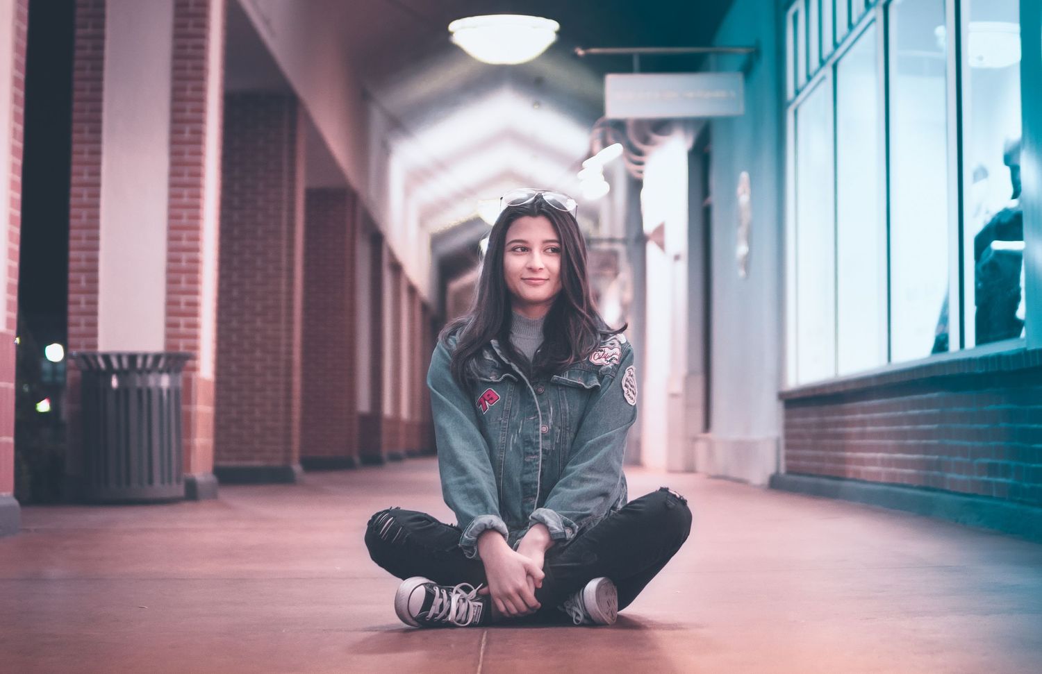 Portrait of a young woman sitting cross-legged in a softly lit corridor, wearing a denim jacket and smiling in an urban night setting