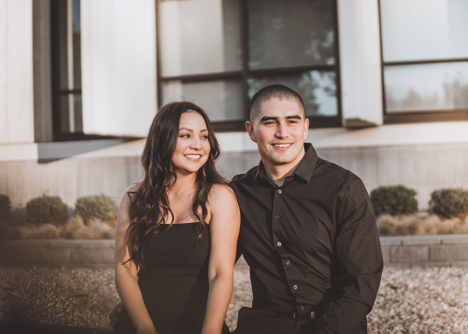 Couple sitting together near a building, smiling in warm golden hour light with a calm, minimal aesthetic and relaxed outdoor atmosphere