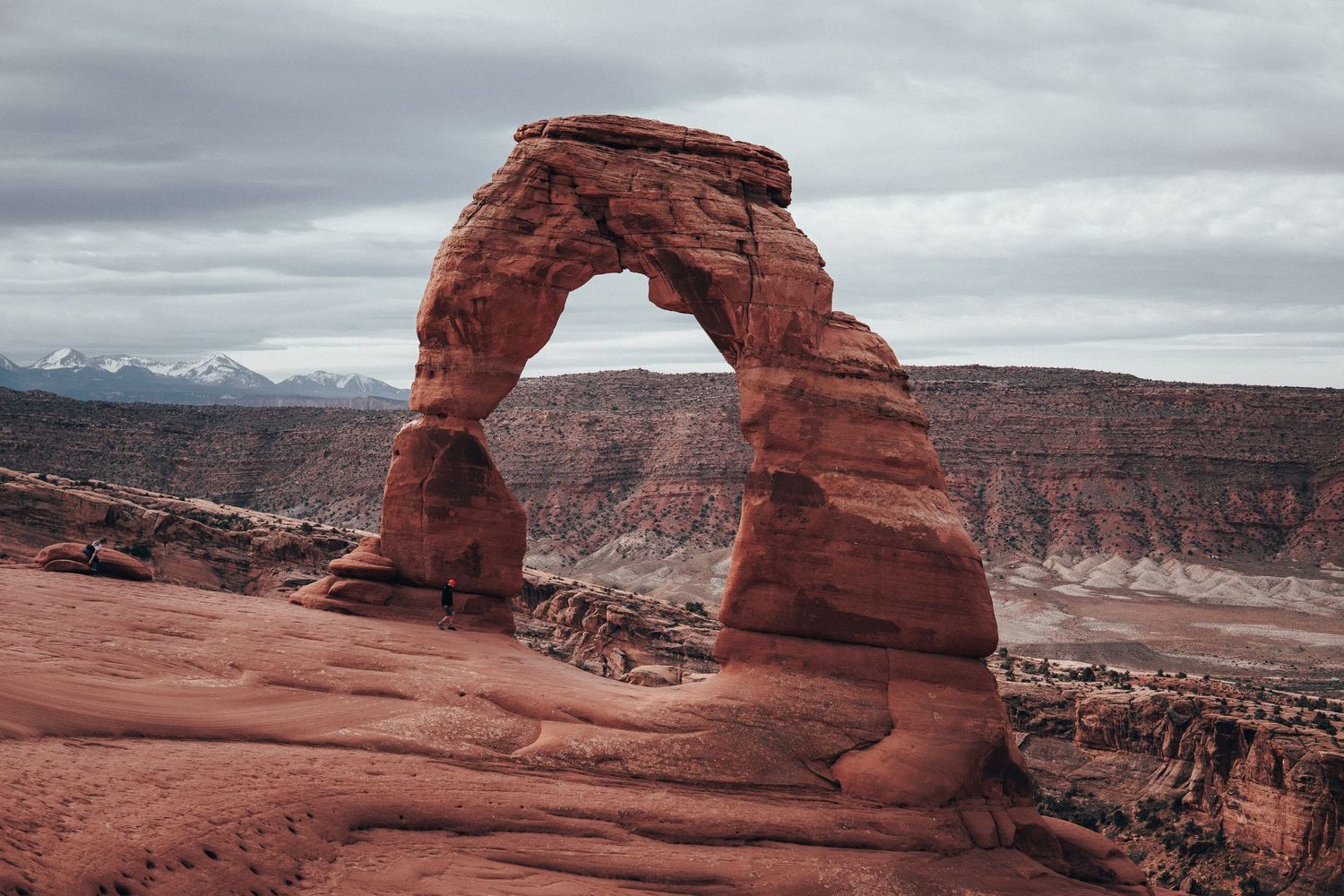 Delicate Arch towering over the red desert landscape in Arches National Park, with hikers nearby for scale and snow-capped mountains visible