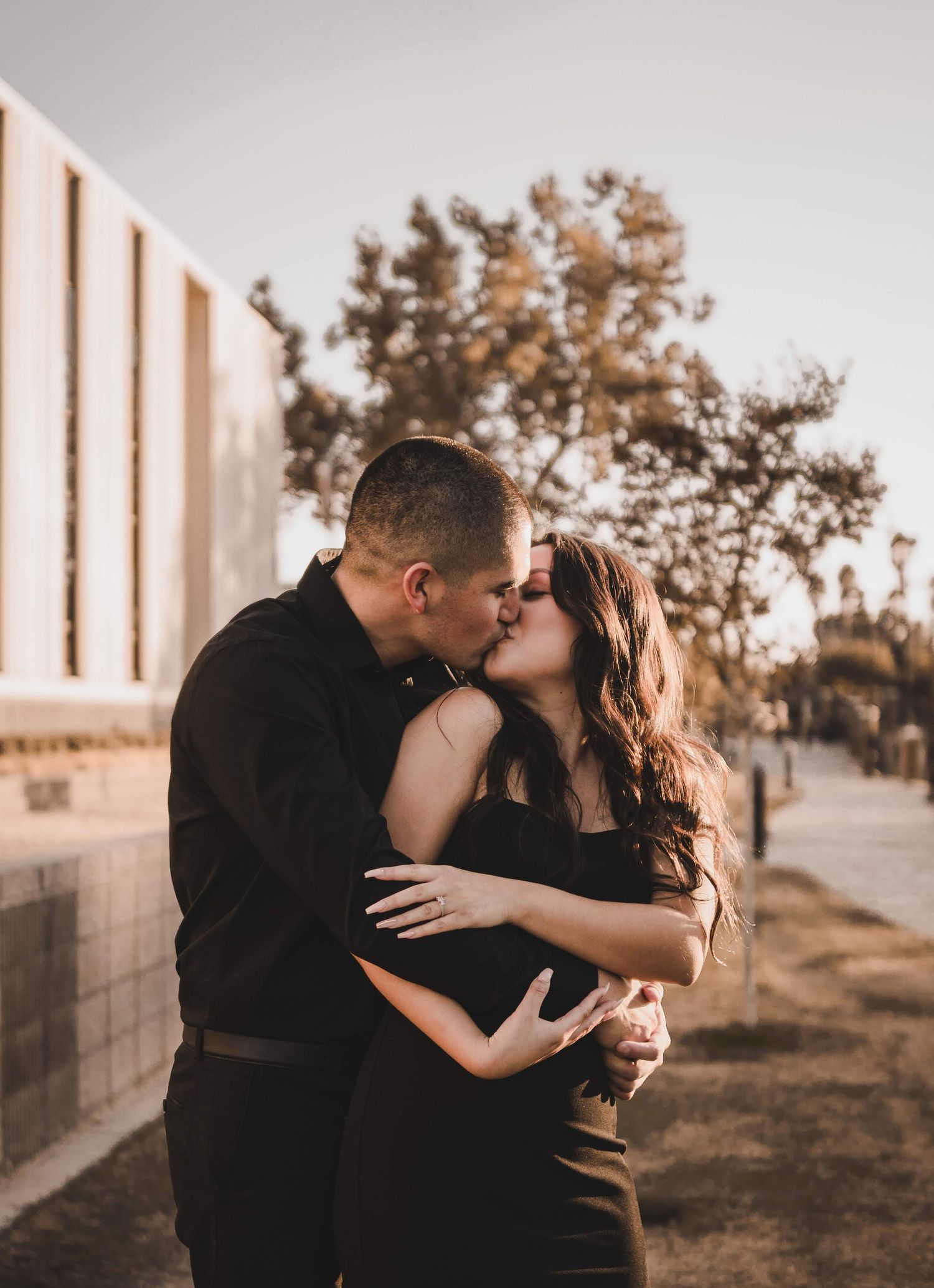 Couple sharing a kiss beside a modern building at golden hour, standing close as warm sunlight highlights their elegant black outfits