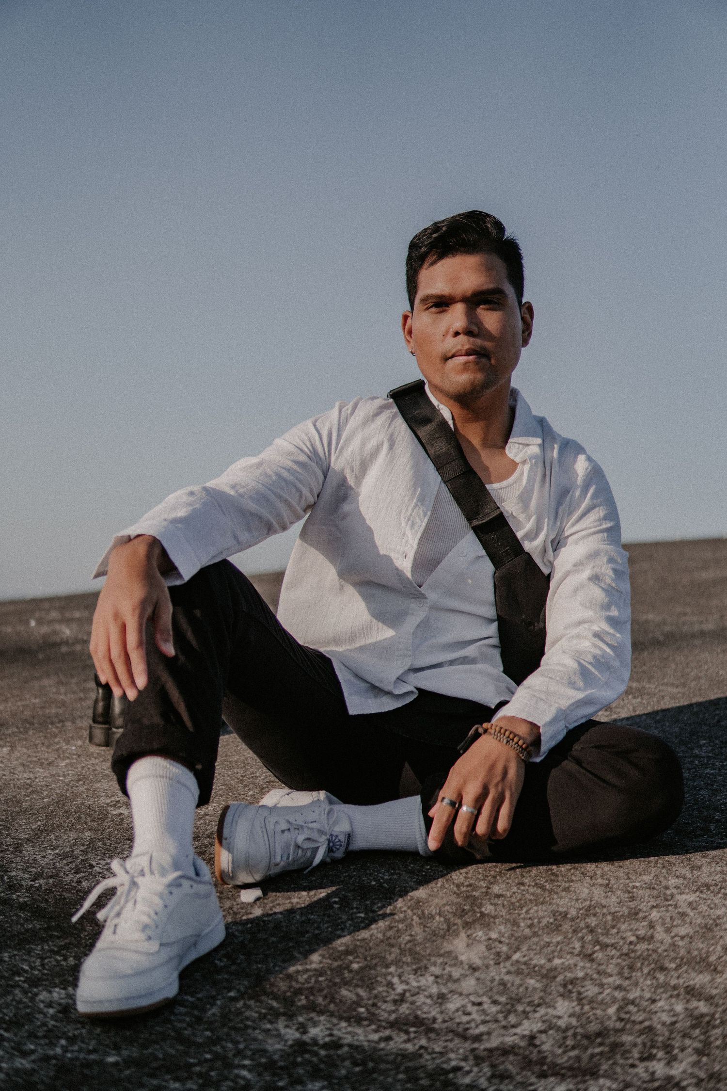 Man sitting casually on a concrete surface in sunlight, wearing a white shirt, black pants, and white sneakers