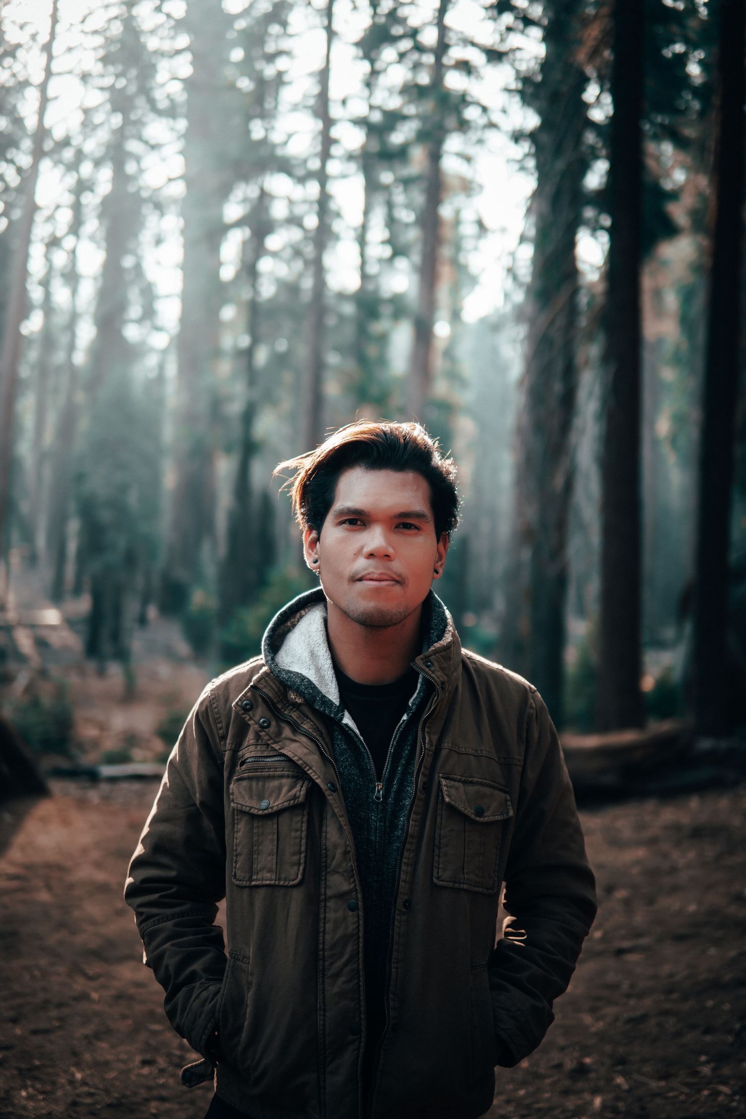 Man in a forest wearing layered jackets, standing in soft morning light among tall trees.