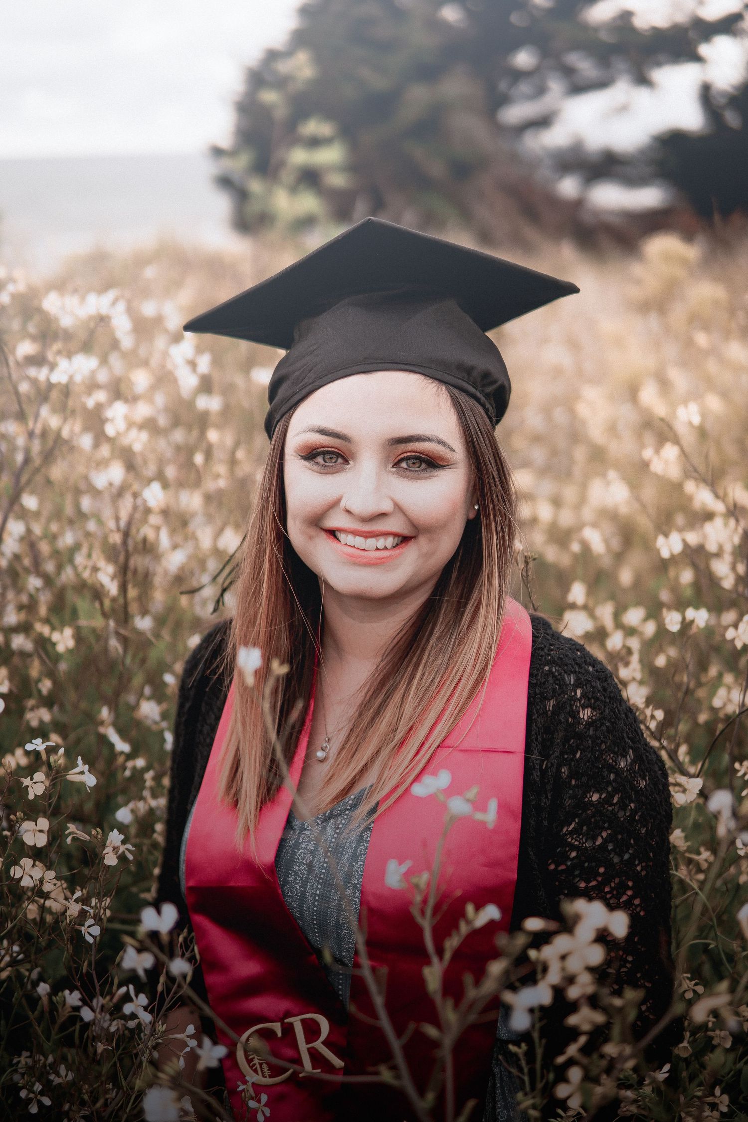 Soft graduation portrait of a young woman in cap and gown smiling in a wildflower field, blending minimal light, nature, and celebration