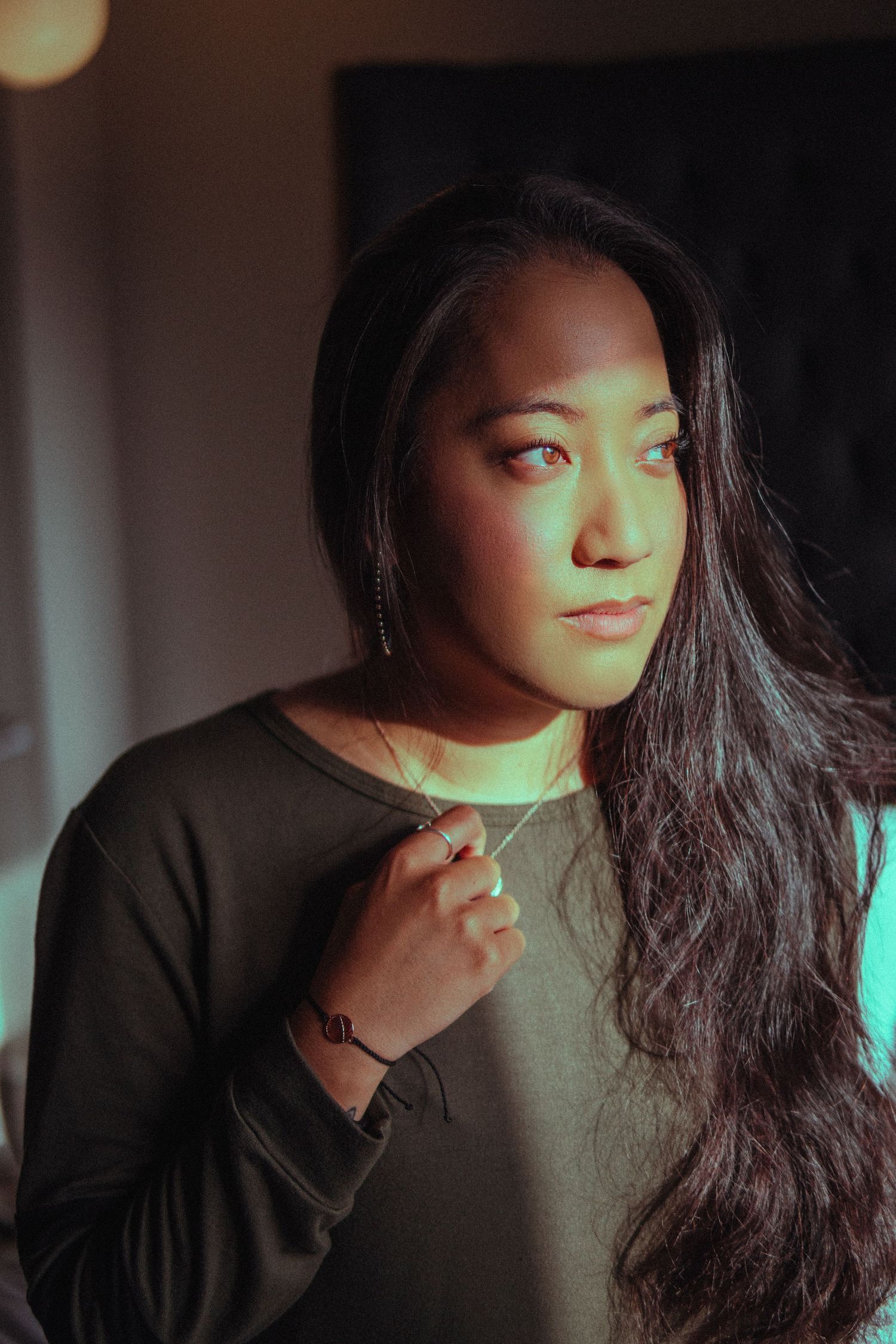 Woman standing in warm window light, gently holding her necklace as sunlight highlights her thoughtful expression and long flowing hair.