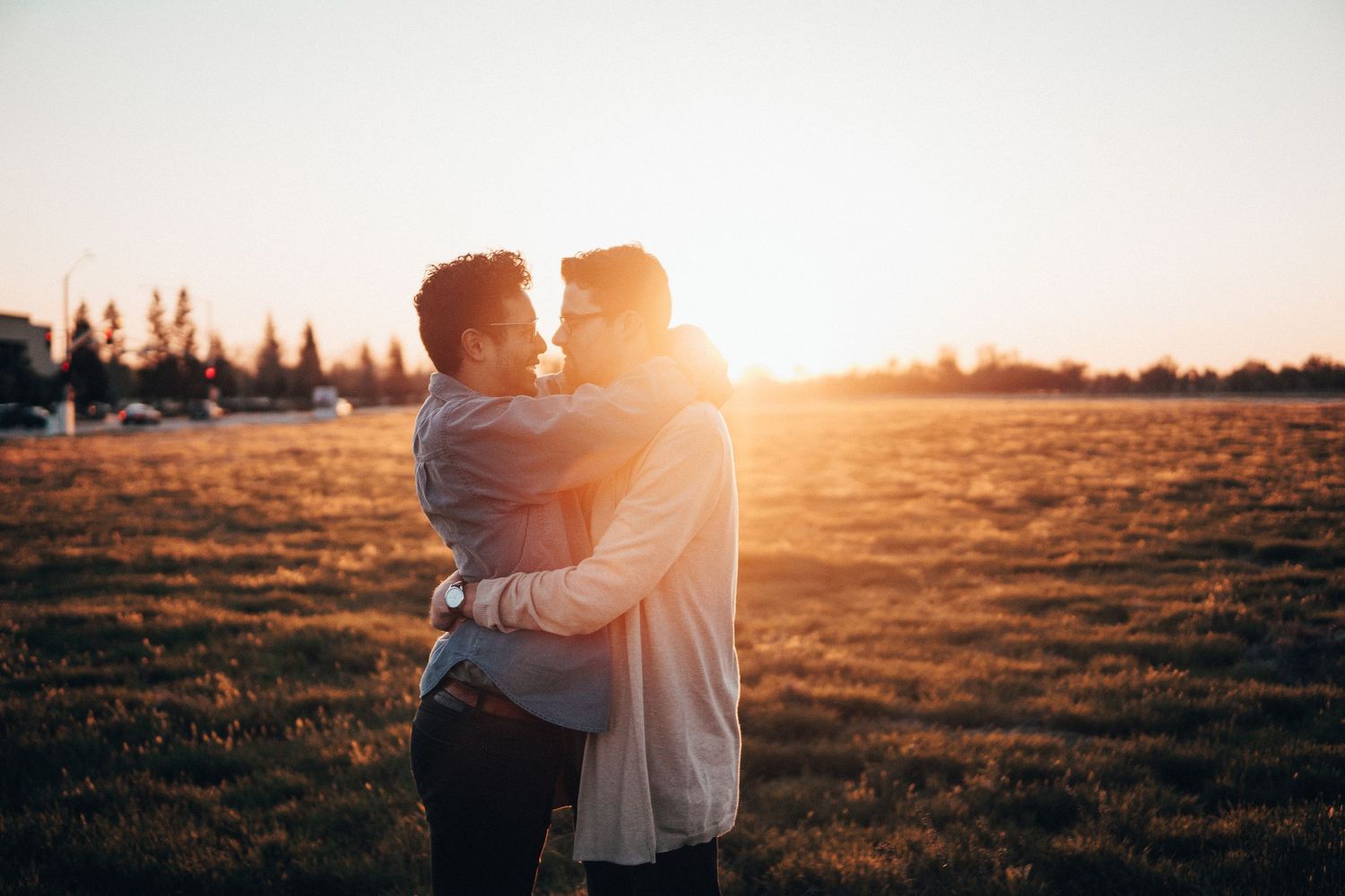 Two people embracing in a sunlit field at golden hour, standing in warm natural light with an open grassy landscape behind them.