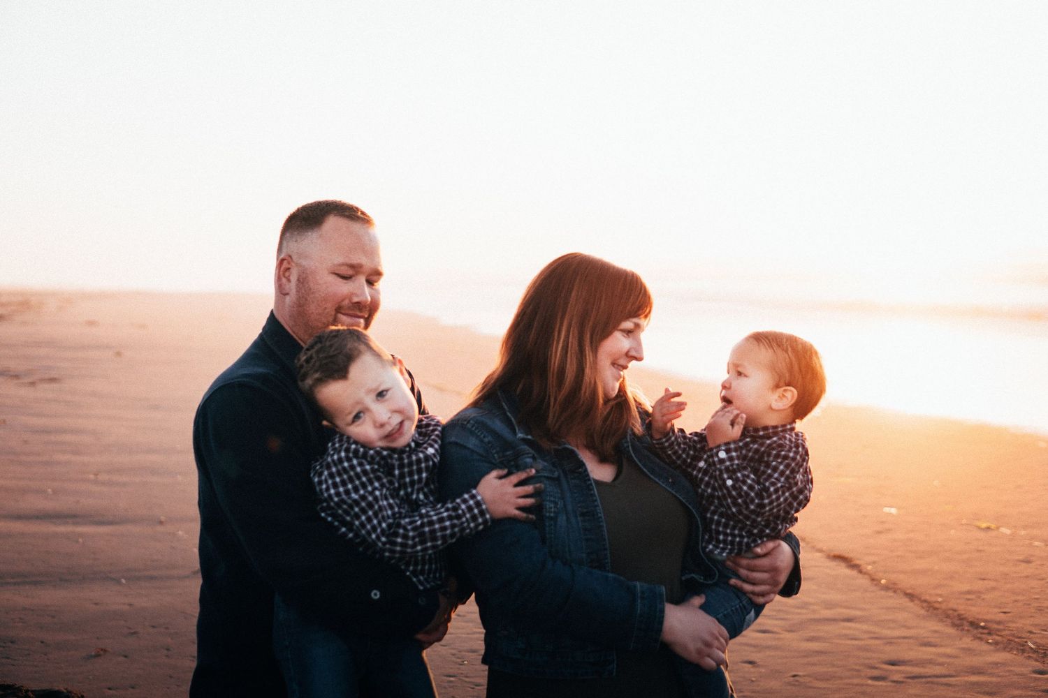 Family holding their young children on the beach at sunset, sharing laughter and warm golden light for a joyful, natural coastal portrait