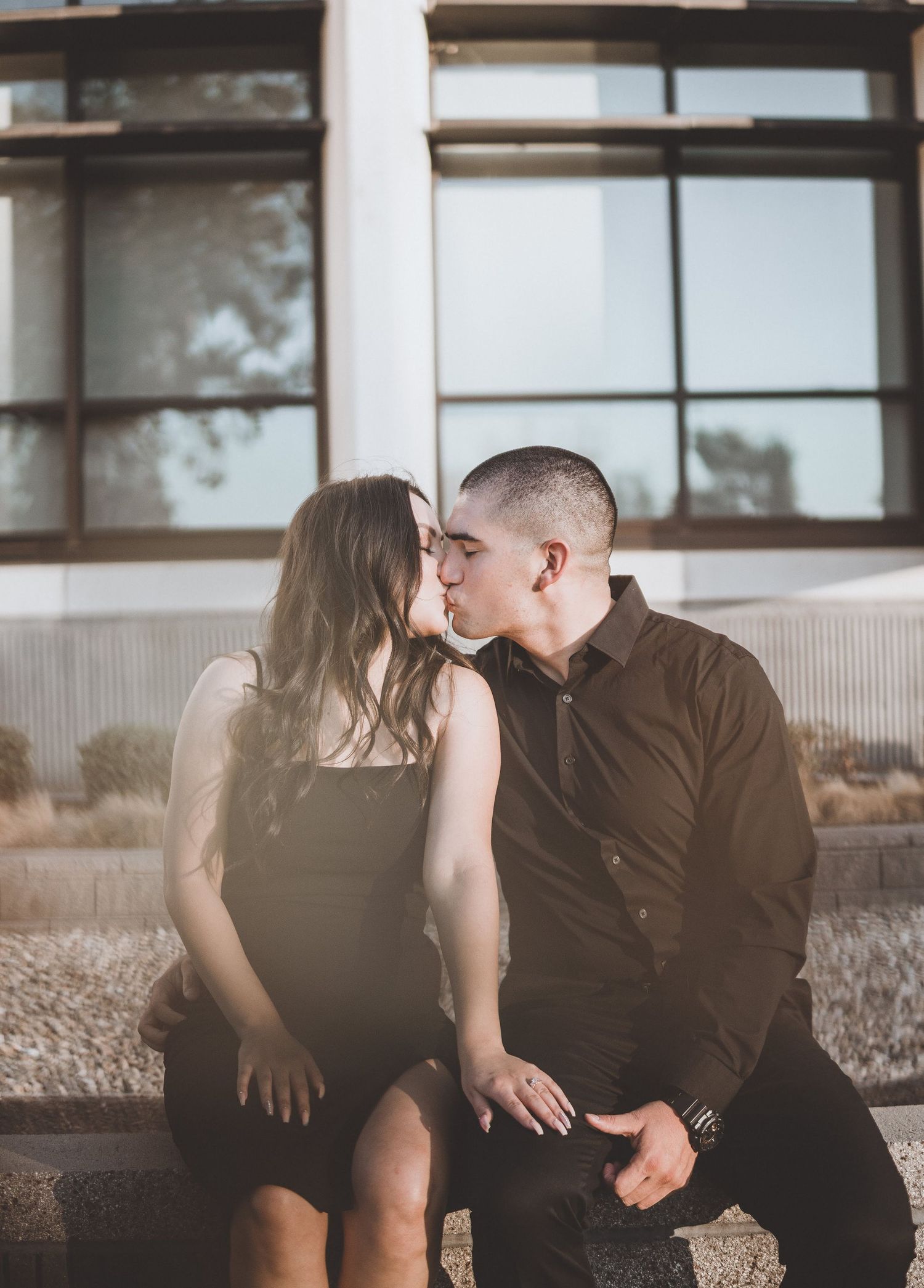 Couple sitting beside a modern building, smiling warmly at each other in soft golden hour light for a relaxed, natural and romantic portrait