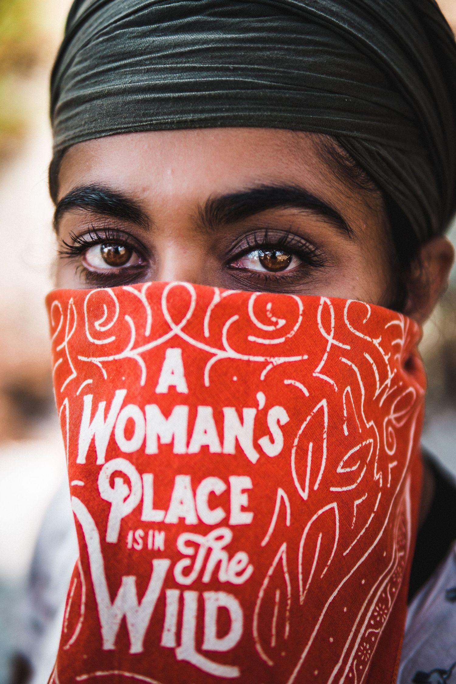 Close-up of a woman with expressive eyes wearing a bandana that reads ‘A Woman’s Place Is in the Wild’ during an outdoor adventure