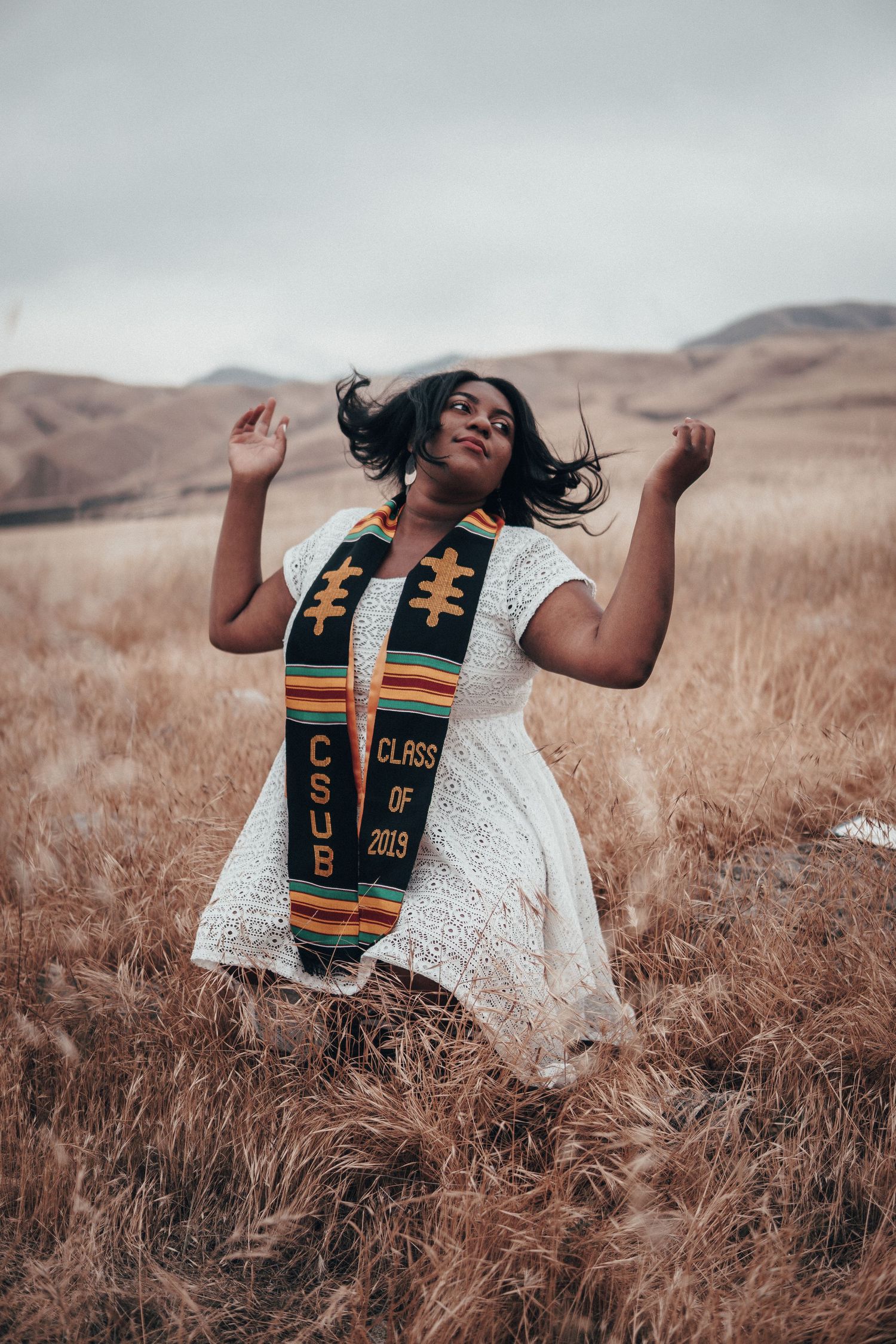 A graduate wearing a white lace dress stole poses confidently in a golden field with rolling hills in the background.