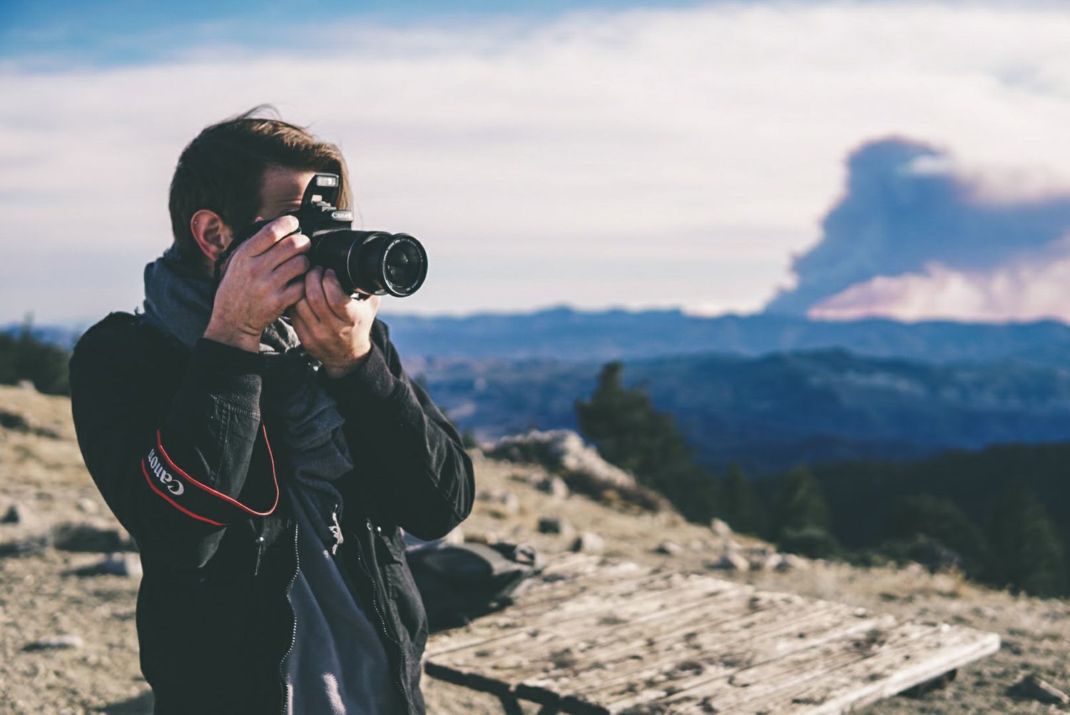 Photographer holding a camera on a windy mountain overlook, capturing views of distant ridges and a dramatic cloud formation.